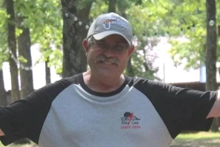 Man with glasses, mustache, and gray hair wearing a beige baseball cap and a blue polo shirt with a logo that says 'Rock Haven Ministries, Heber, Arkansas', standing against a plain white wall.