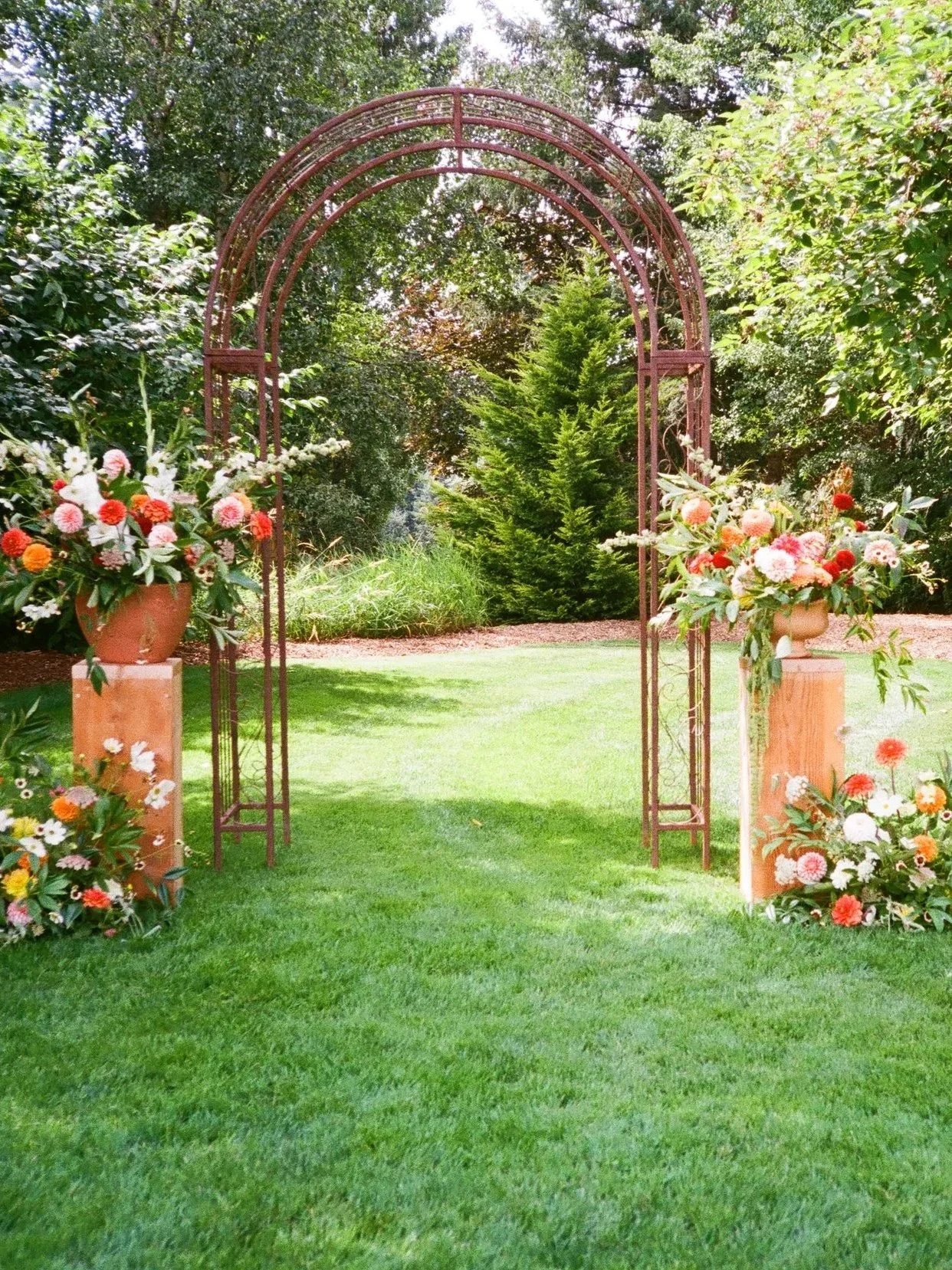 A decorated outdoor garden arch with floral arrangements on two pedestal stands on either side of the arch, set on a lush green lawn.