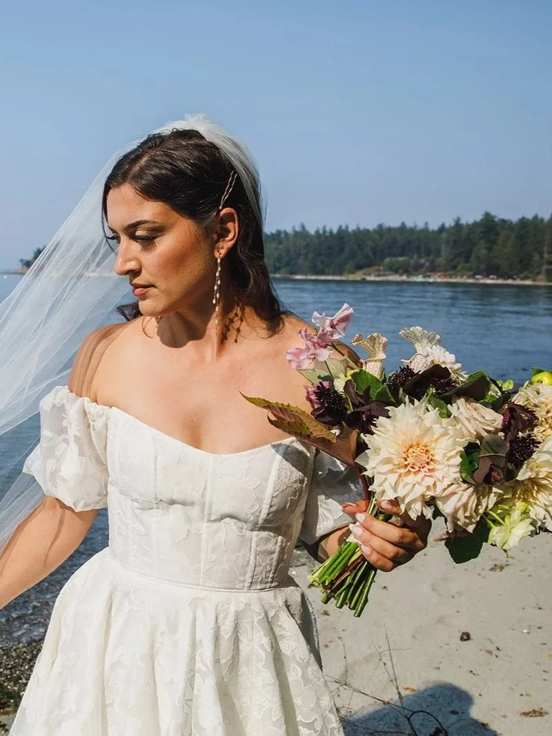 A bride in a white off-shoulder wedding dress stands on a beach holding a bouquet of flowers, with the ocean and a distant shoreline with trees in the background.