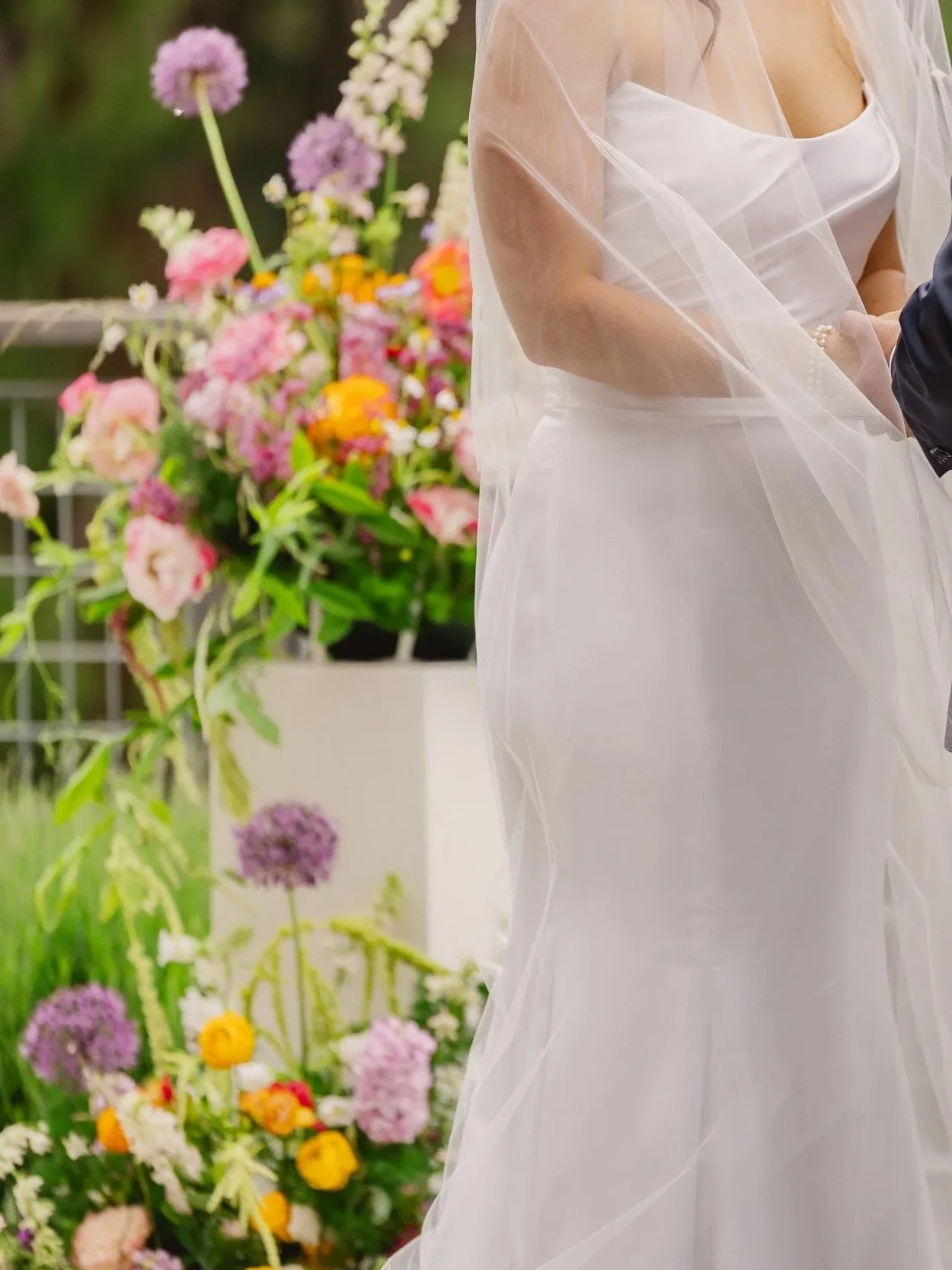 A bride and groom holding hands during their wedding ceremony outdoors with colorful flowers in the background.