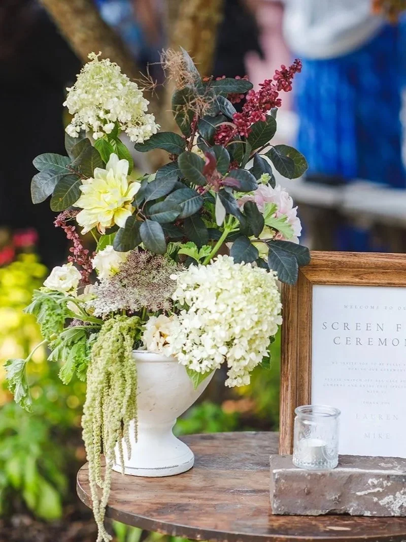 A wedding sign reading 'Screen Free Ceremony' in a wooden frame, next to a white vase filled with white, pink, and purple flowers, and a small candle holder on a brick platform, outdoors with guests in the background.