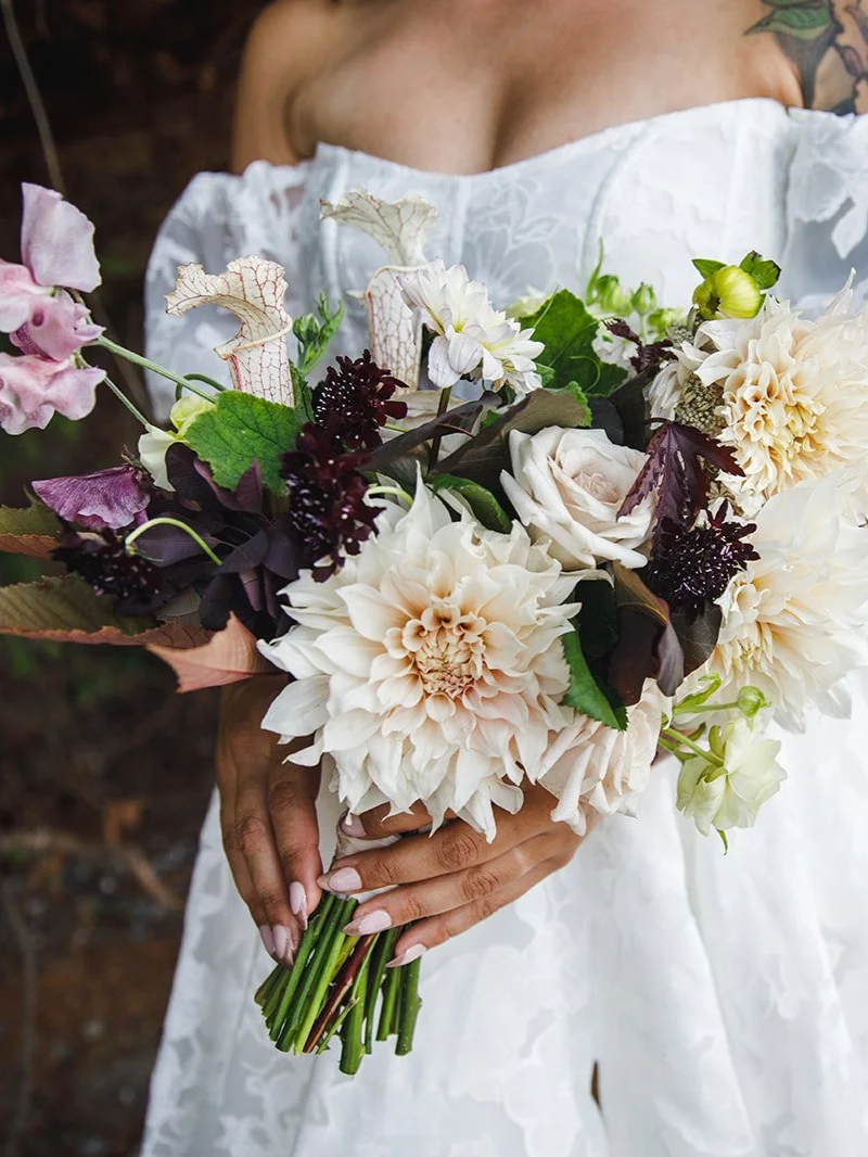 A woman in a white dress holding a large bouquet of white, cream, and dark-colored flowers with green leaves.