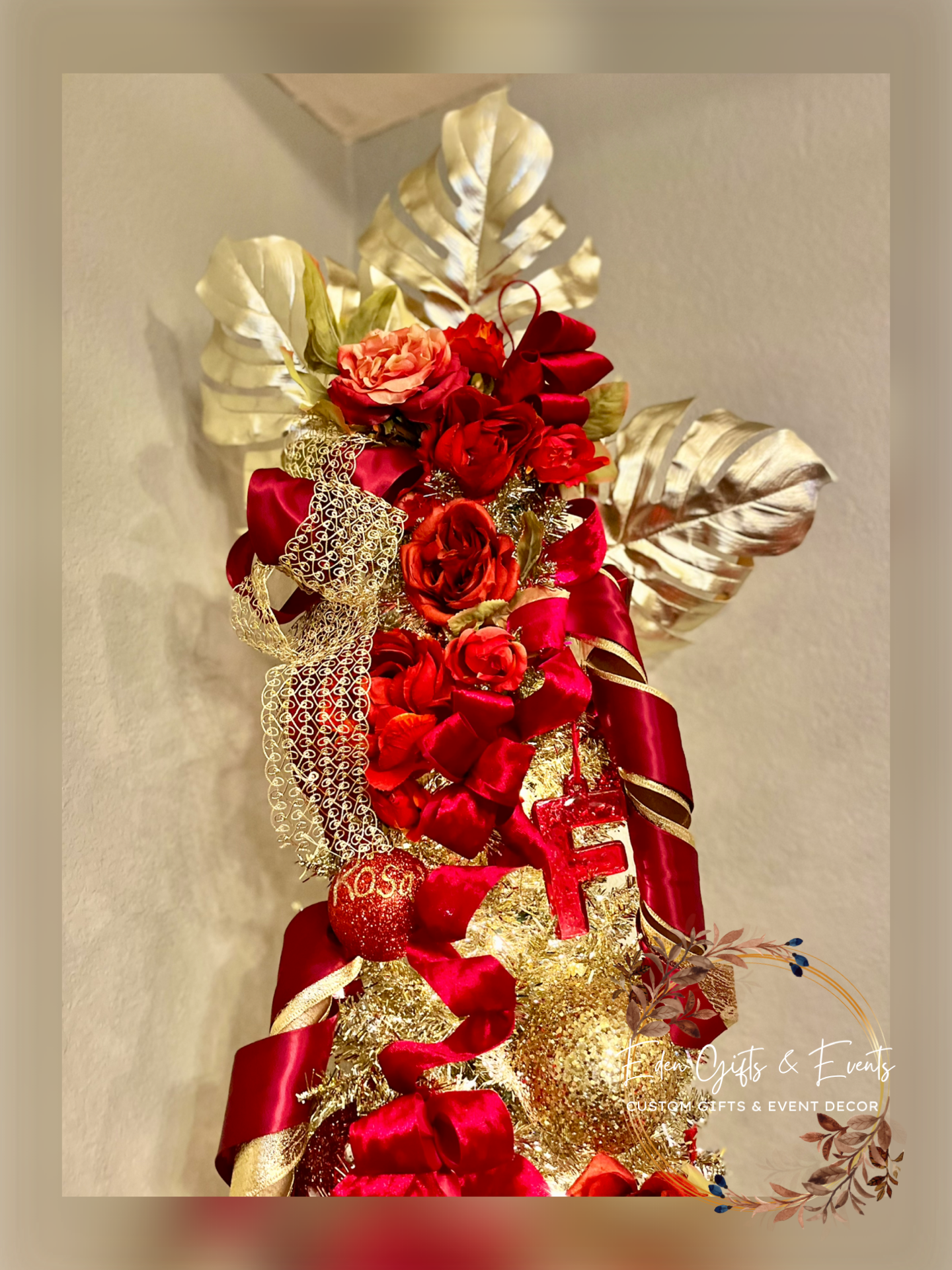 A festive Christmas floral arrangement with red roses, gold leaves, red ribbons, and holiday decorations on a wall.