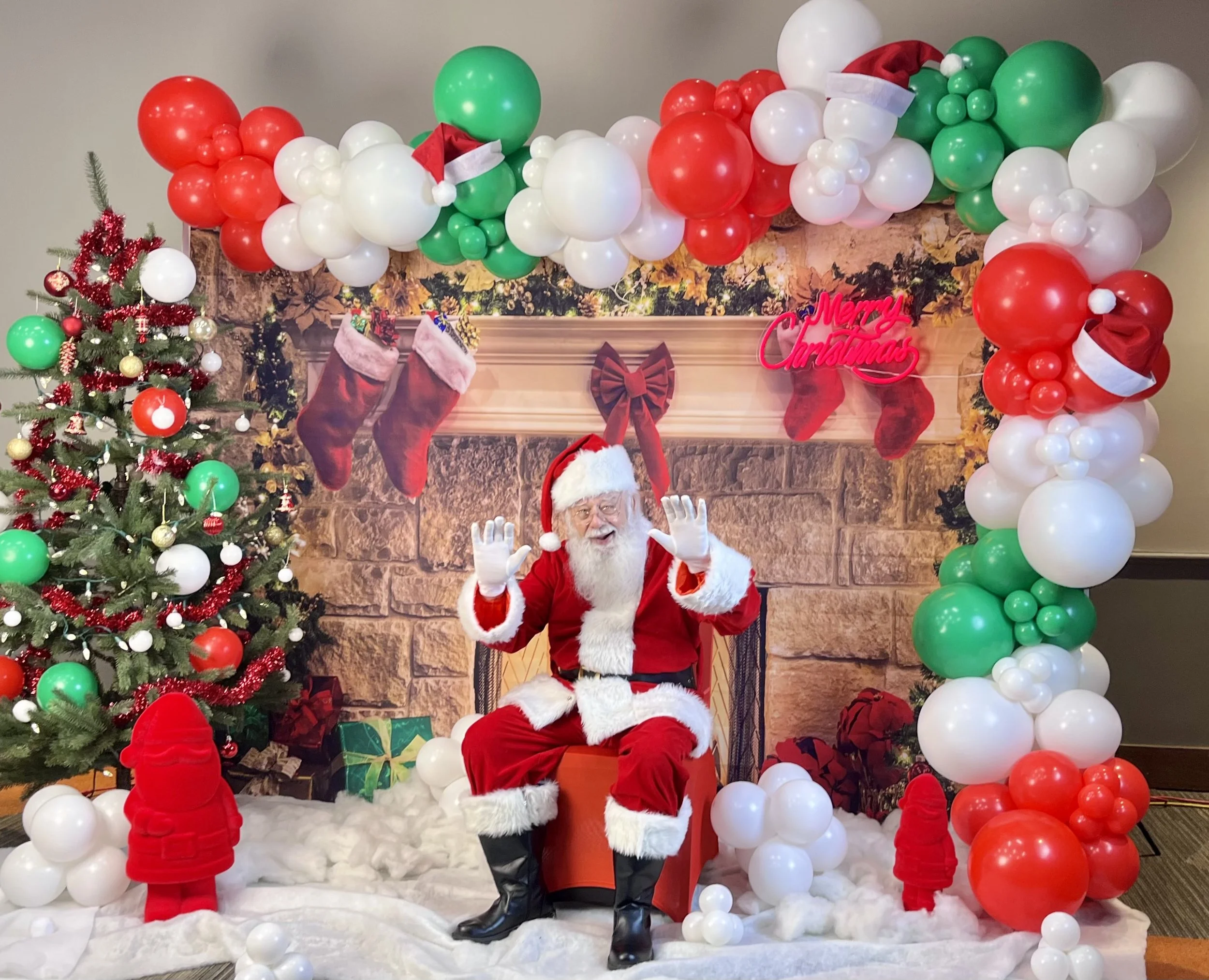 Santa Claus sitting in a decorated Christmas scene with a fireplace backdrop, Christmas tree, balloons, and holiday decorations.