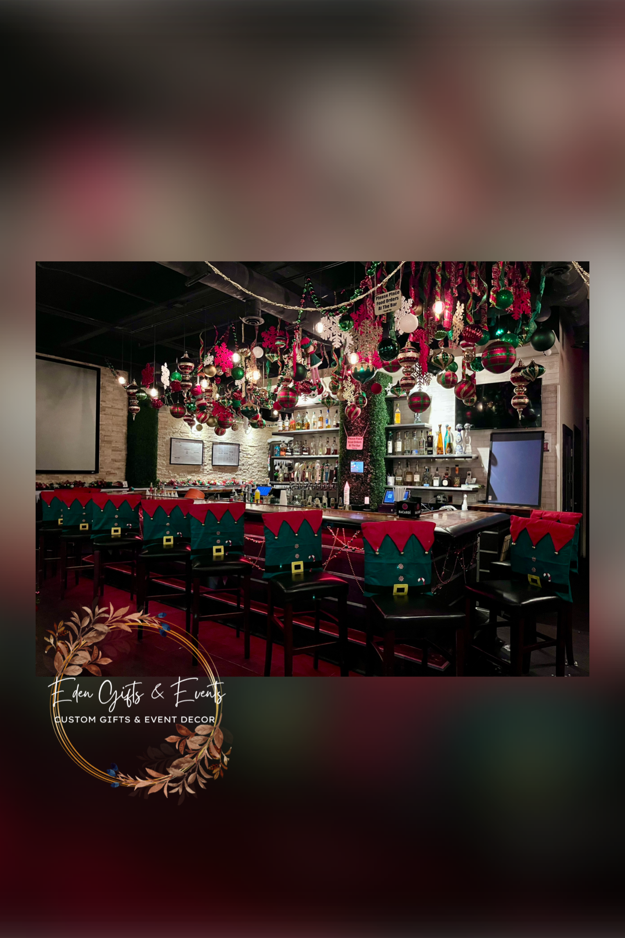 Bar decorated with Christmas ornaments, including hanging baubles, garlands, and elf hat-themed seat covers on barstools, with a sign that reads 'Eden Gifts & Events Custom Gifts & Event Decor' at the bottom.