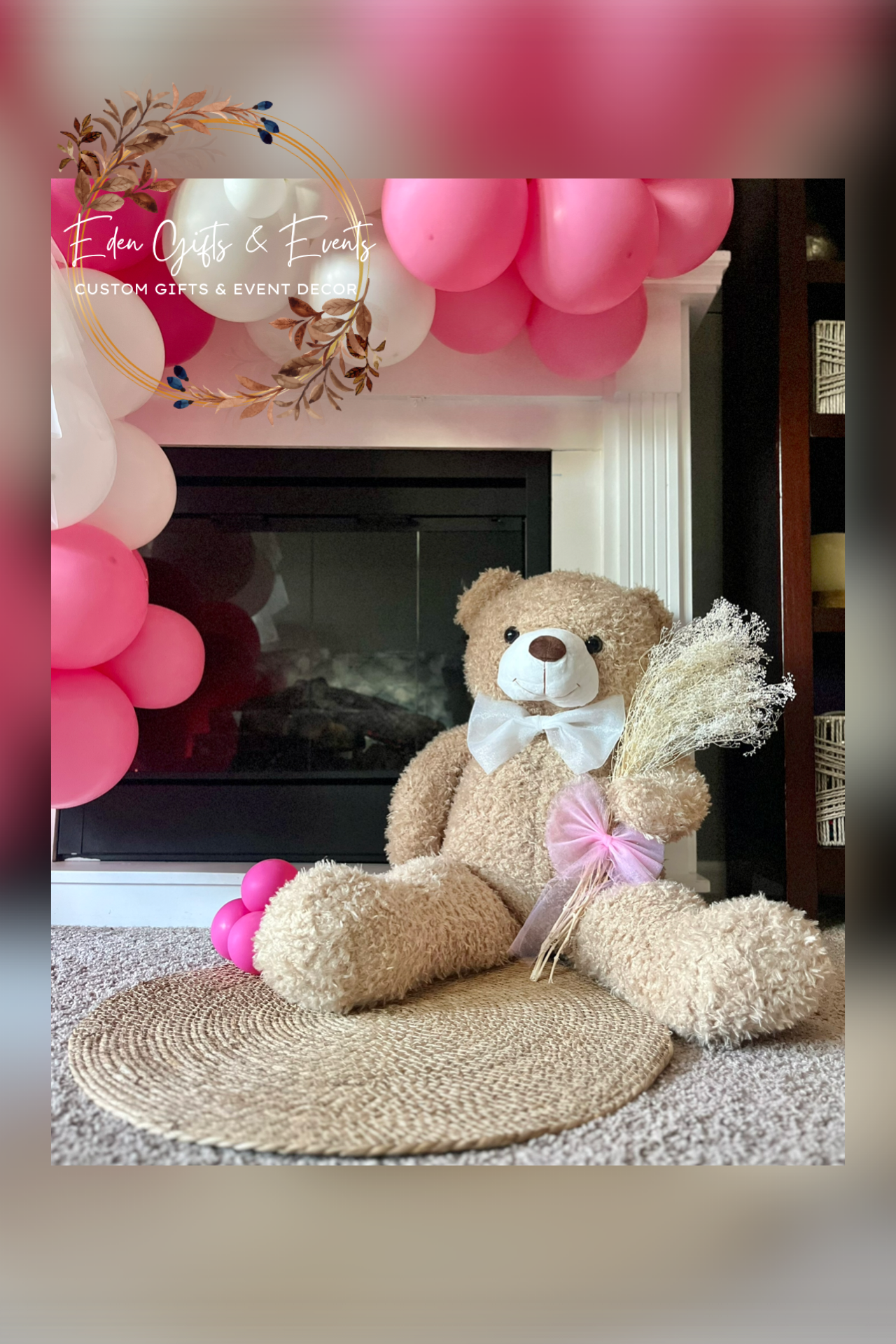 A teddy bear with a white bow tie sitting on a round woven mat in front of a fireplace, surrounded by pink and white balloons and holding a bouquet of dried flowers.