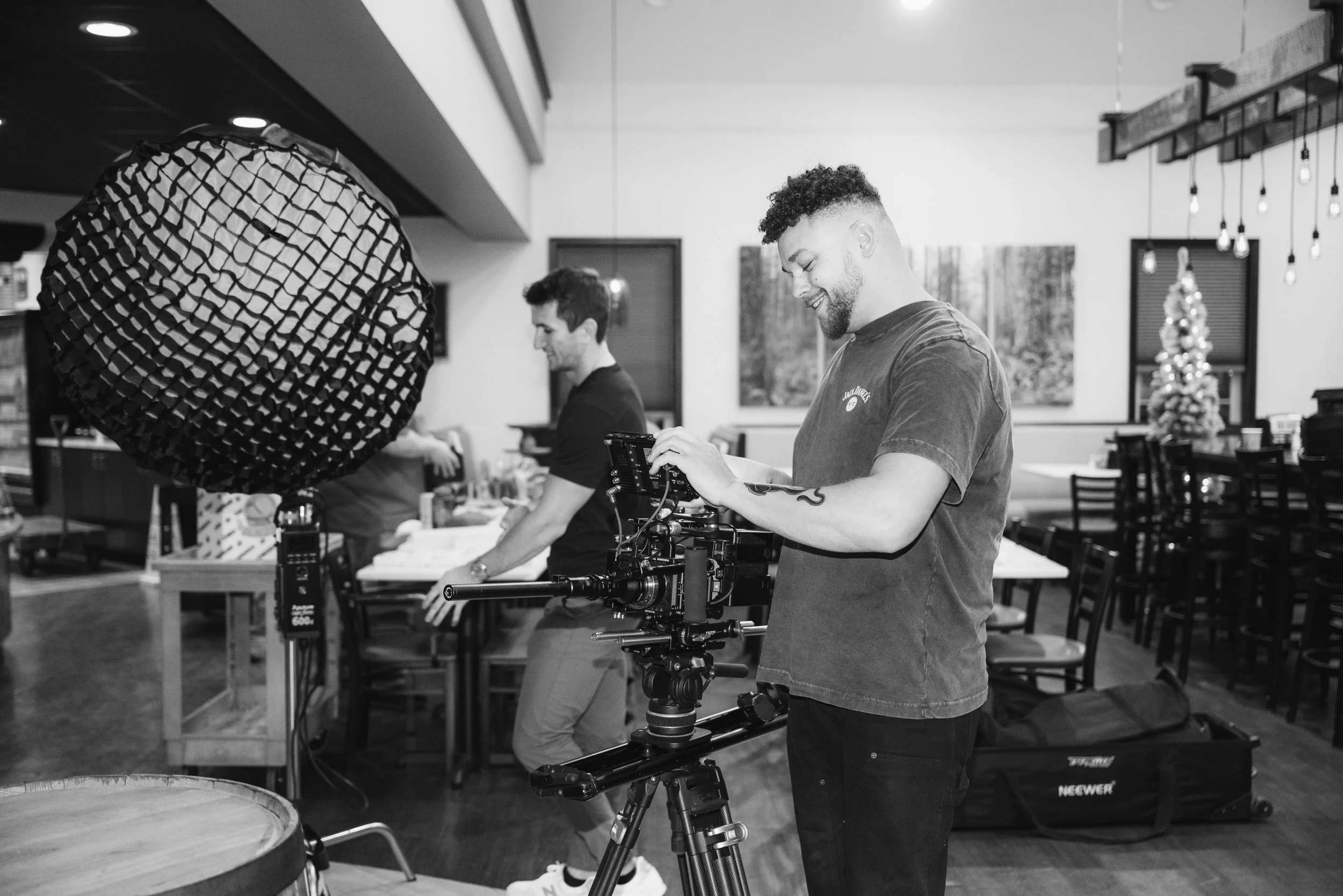 Two men are setting up filming equipment in a decorated restaurant. One man is adjusting a camera on a tripod, and the other is handling a large softbox light. There is a Christmas tree in the background.
