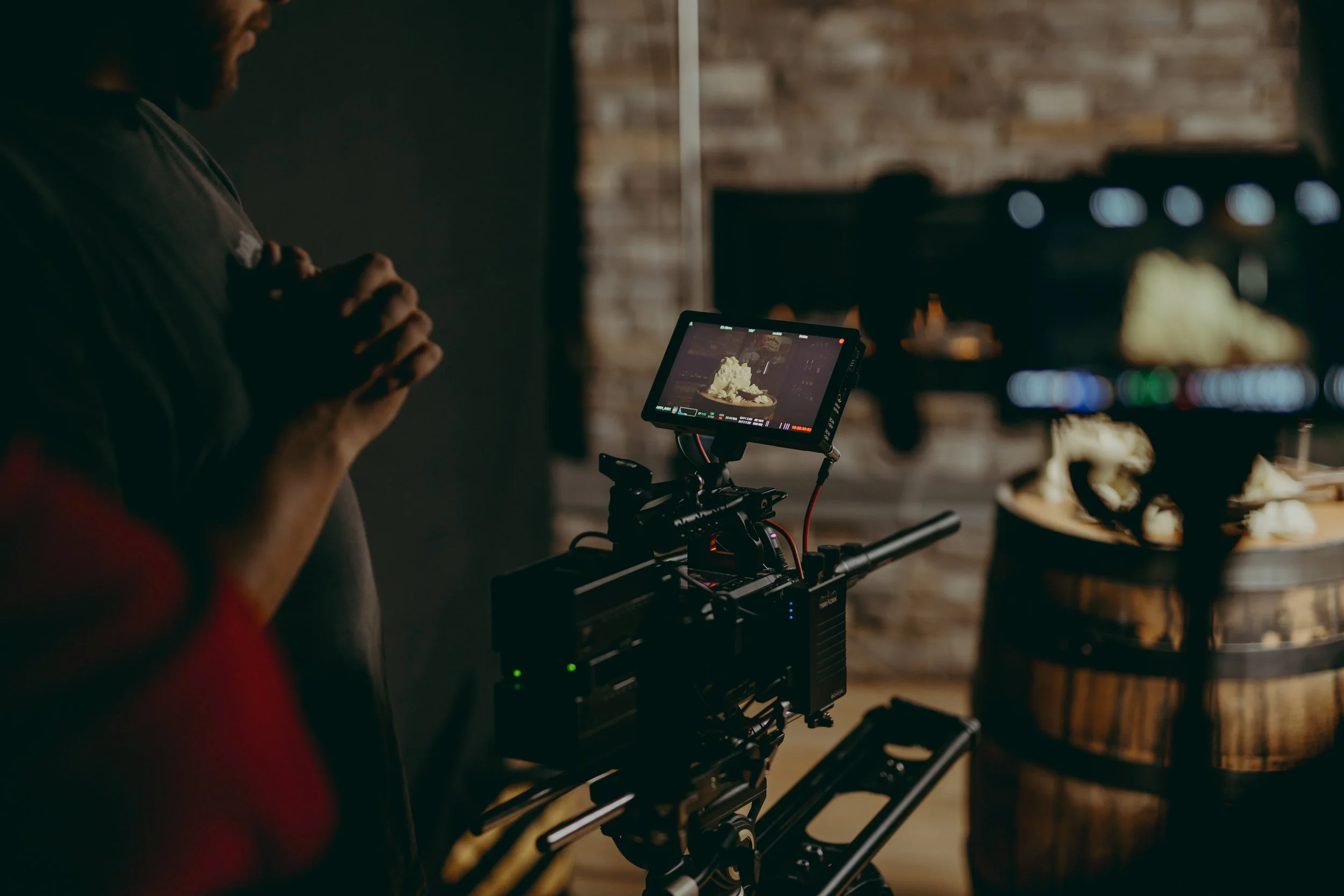 A person operating a camera in a dimly lit room with a brick wall, focusing on a food shoot setup centered on a wooden barrel with food items inside.