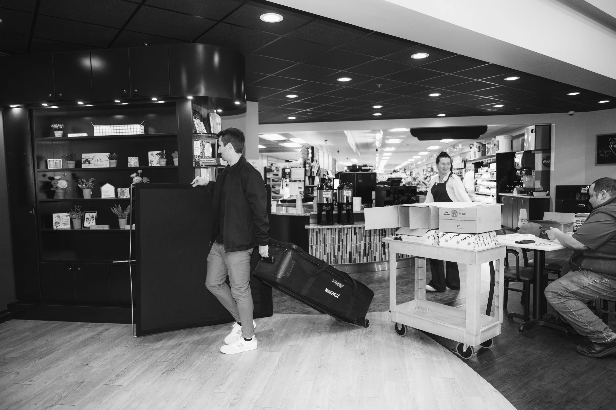 A man pulling a rolling suitcase past a counter in a store or cafe, with a woman behind the counter and another person seated at a table.