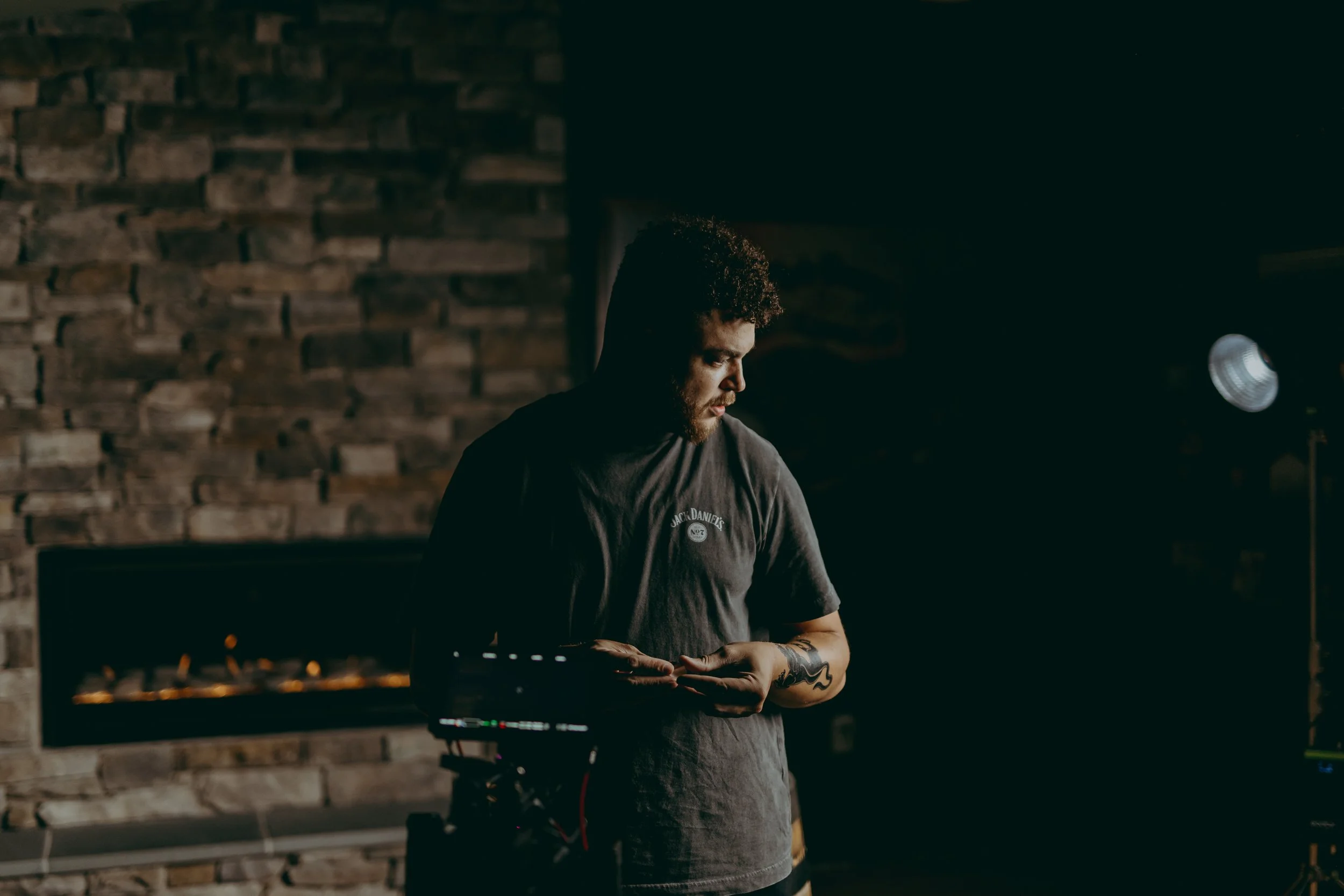 A man with curly hair and a beard standing in a dimly lit room with a brick wall, holding a small object and looking down. There is a fireplace and a studio light in the background.