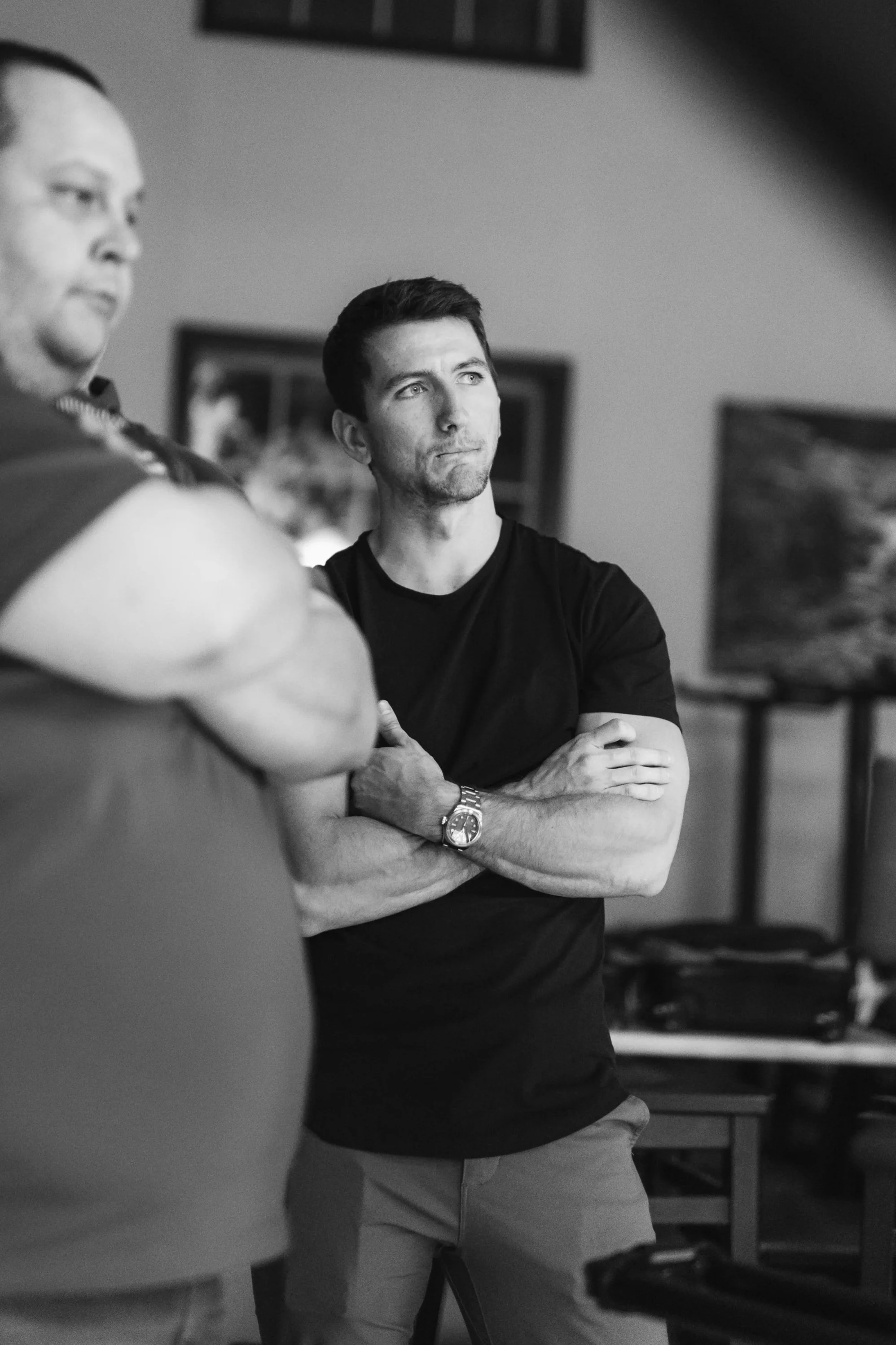 Black and white image of two men in a room, one in the foreground with arms crossed, partially visible, and the other in the background with arms crossed, looking to the side.
