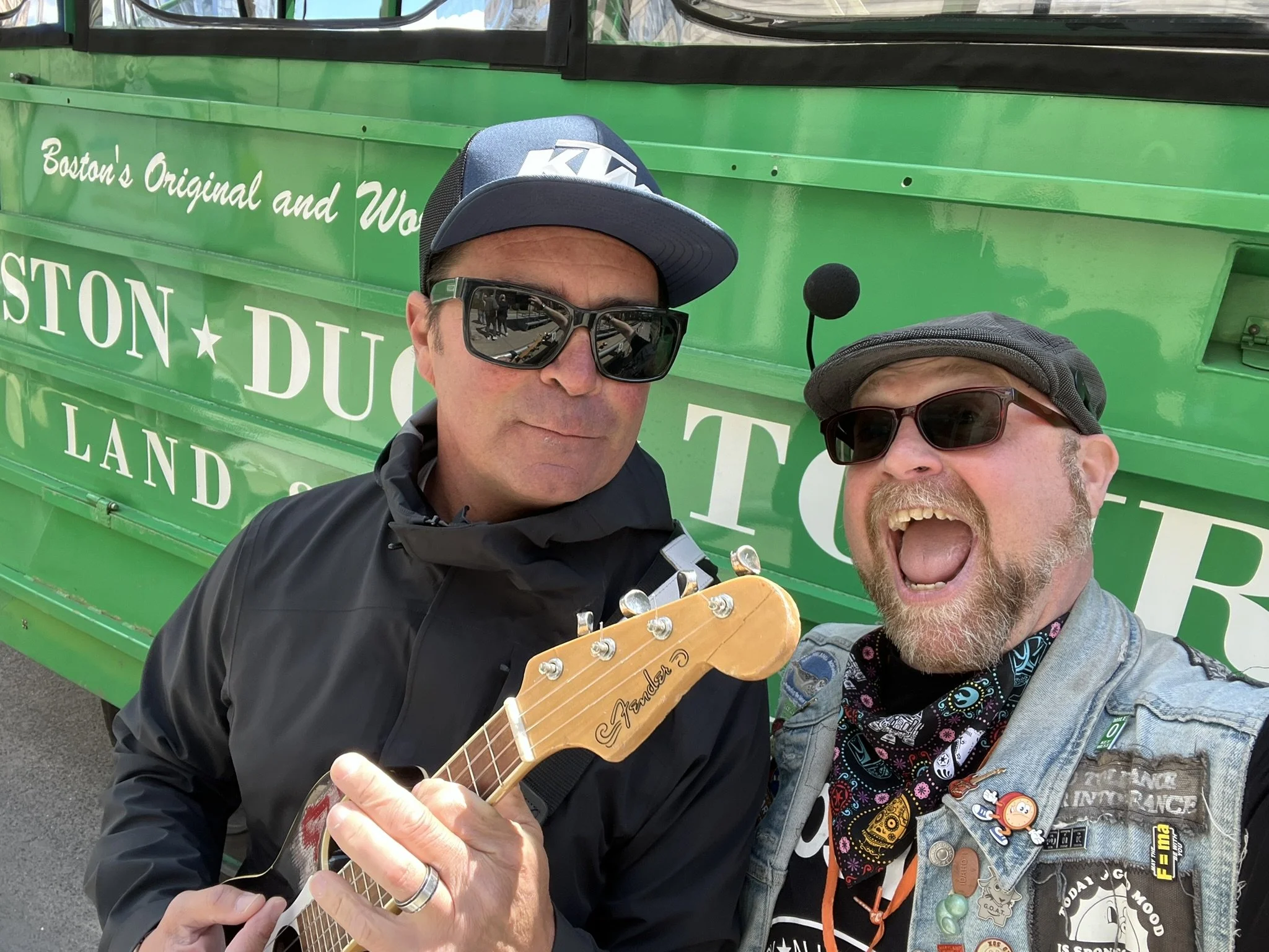 Two men taking a selfie in front of a green vehicle, one man holding a guitar, both wearing sunglasses and casual clothing.