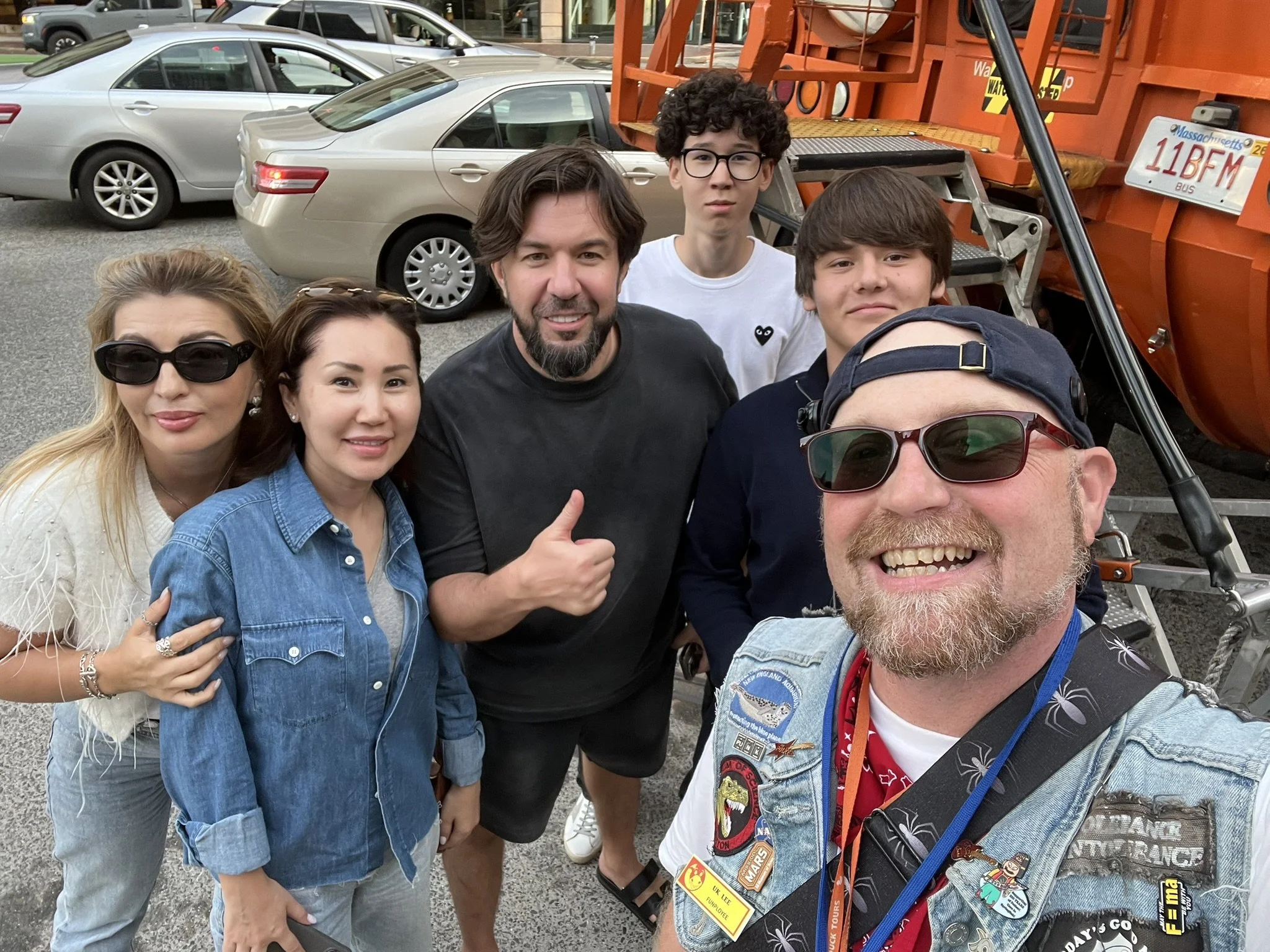 A group of six people taking a selfie with tour guide U.K. Lee outside a DUKW and a street visible in the background. The group consists of three women and three men, some wearing sunglasses and casual clothing, smiling or making gestures.