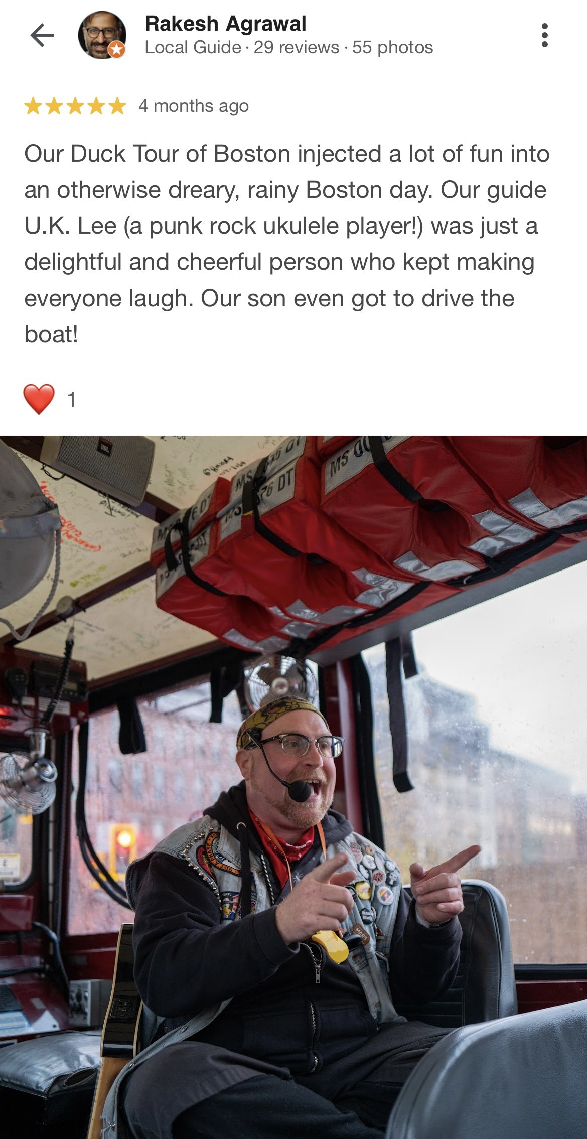 A man with glasses, a headscarf, and a microphone headset sitting inside a bus, smiling and gesturing with his hands. The bus has a red life-saving bag and some electronic equipment visible in the background.