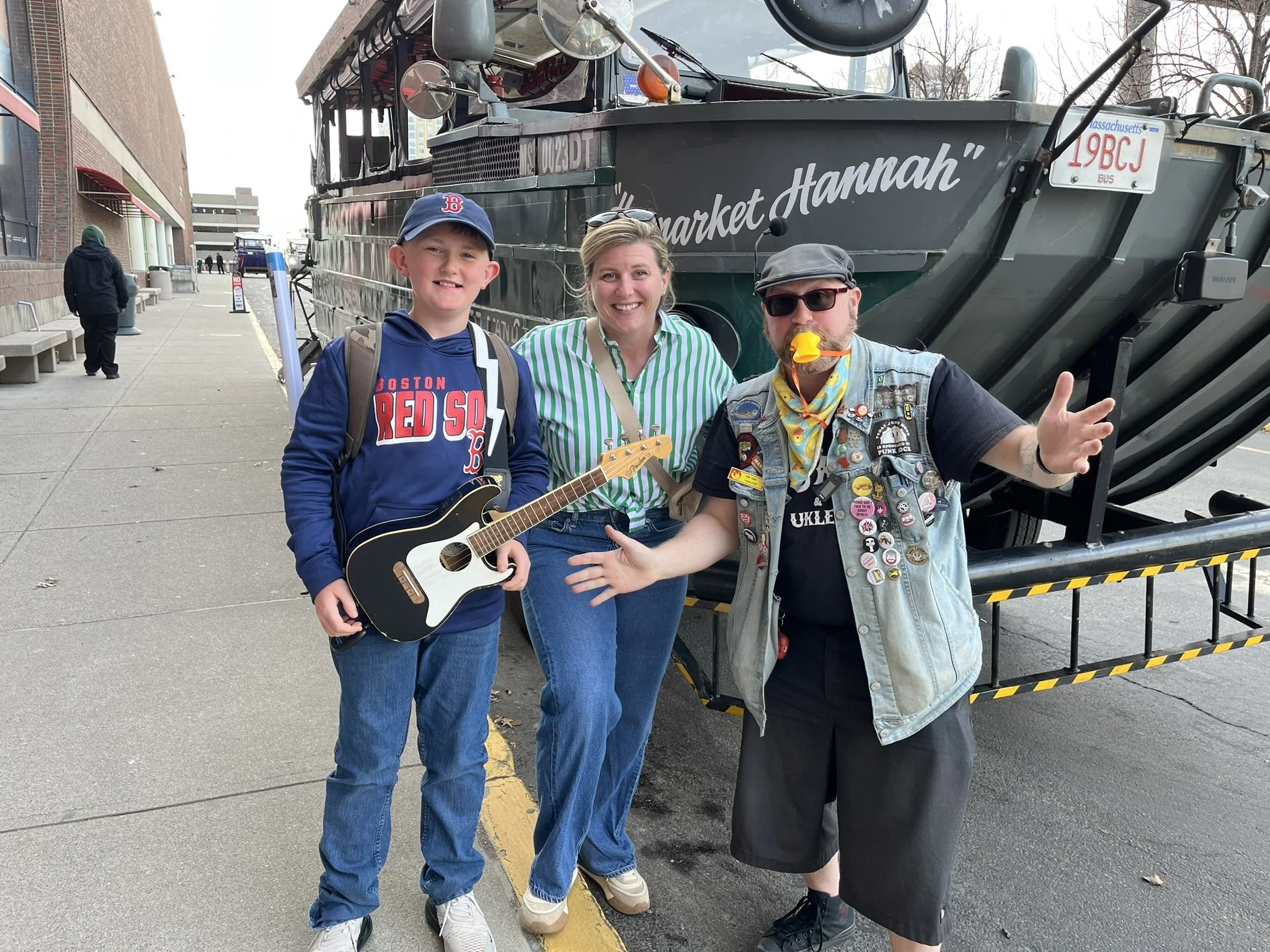 A young boy and two adults standing under a boat with the words "Market Hannah" on it. The boy is wearing a navy blue baseball cap, a blue hoodie with "Boston Red Sox" on it, and holding a toy guitar. The woman is wearing a green and white striped shirt, and the man is wearing sunglasses, a grey cap, and a vest with numerous pins and patches, and has a pacifier in his mouth shaped like a yellow duck. The setting appears to be outdoors on a city sidewalk.