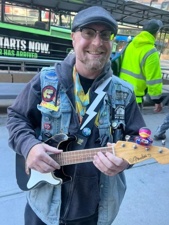 Tour guide U.K wearing glasses, a baseball cap, and a denim vest with patches, holding a Fender electric ukulele, with a replica toy duck figure of himself on the headstock, in an urban outdoor setting with other people in the background.