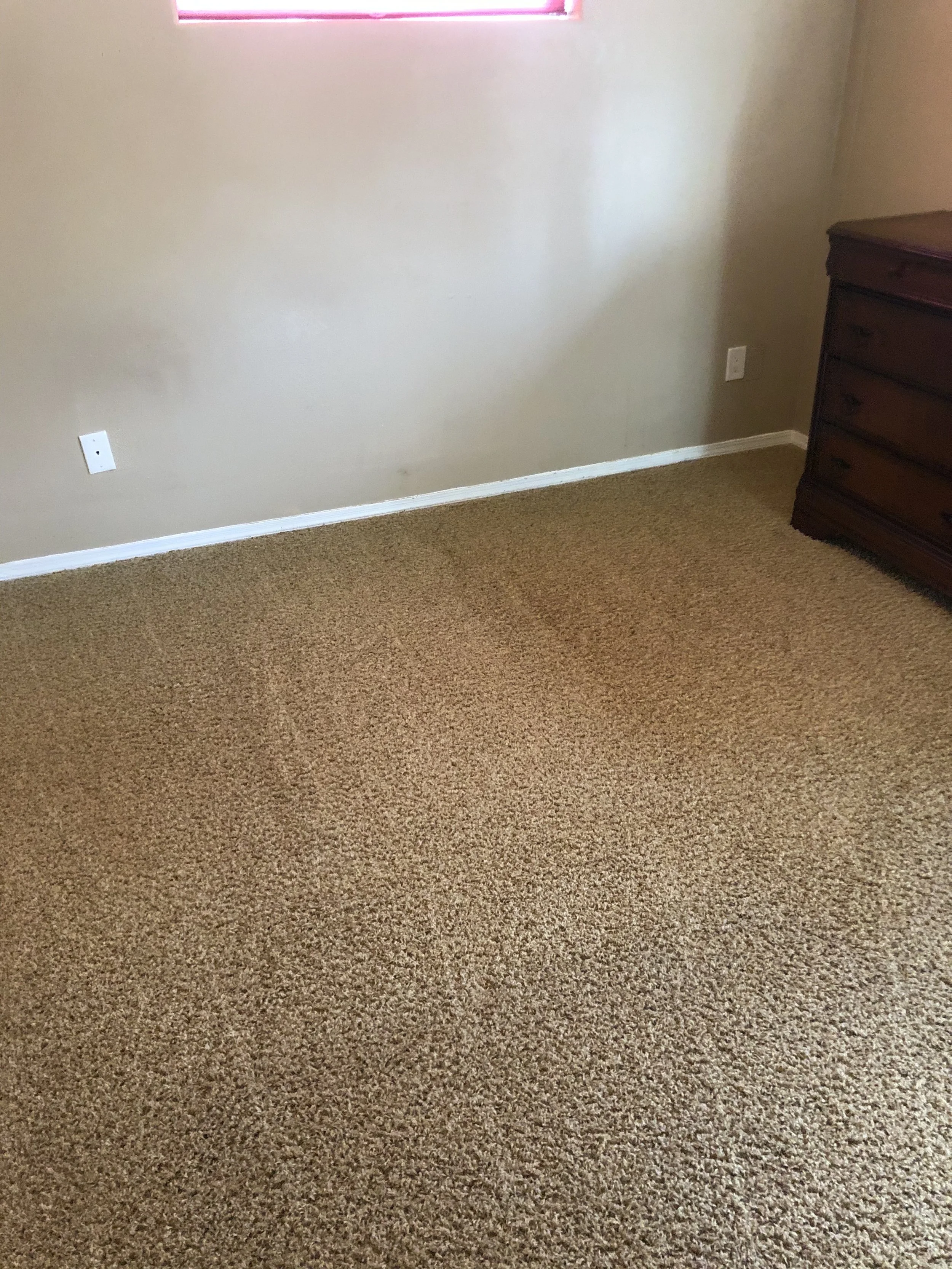 Empty room with beige carpet and walls, a wooden dresser in the corner, and a small window with red curtains.