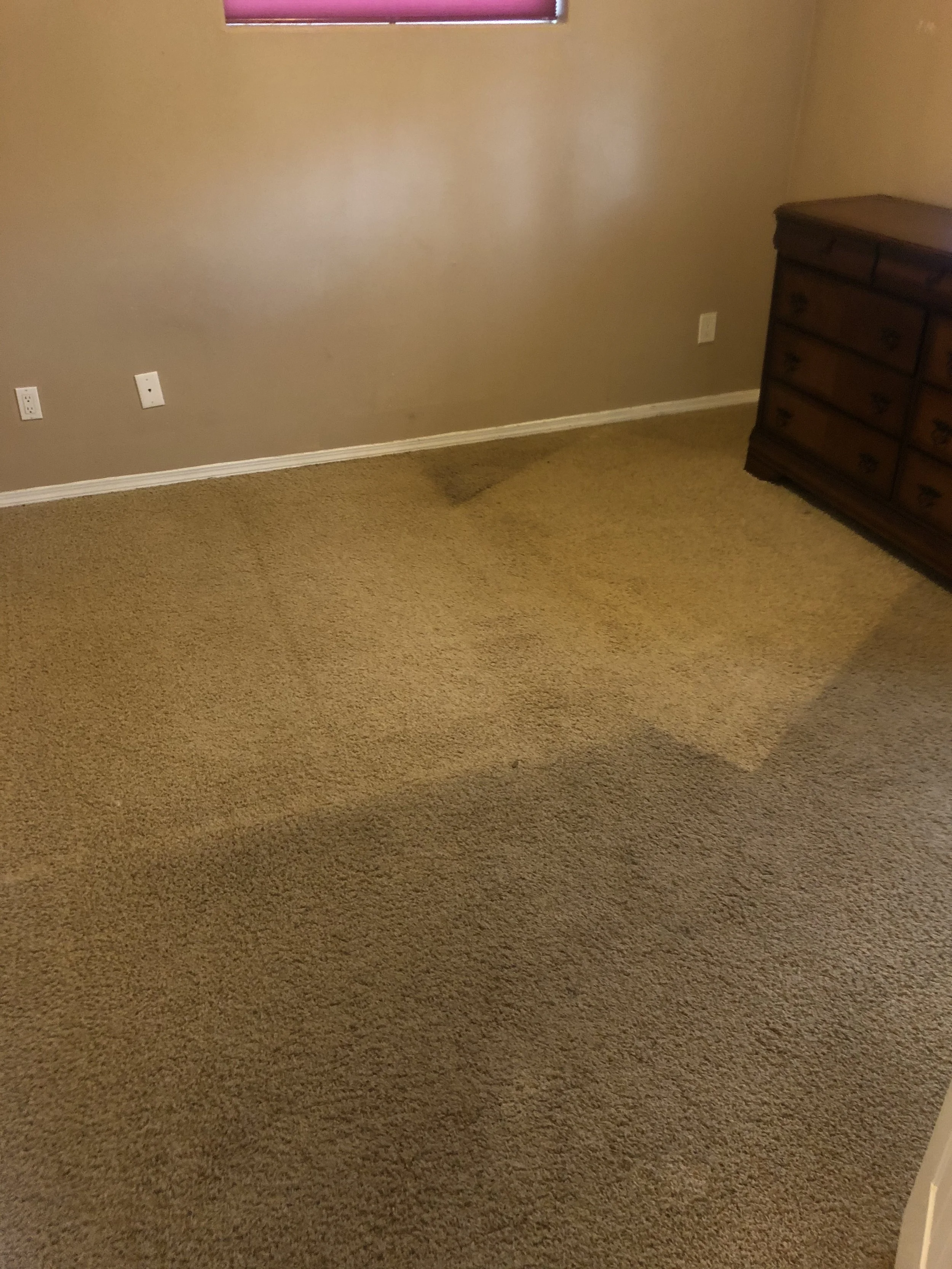 Empty room with beige carpet, wooden dresser, tan walls, and a window with a partially closed red blind.