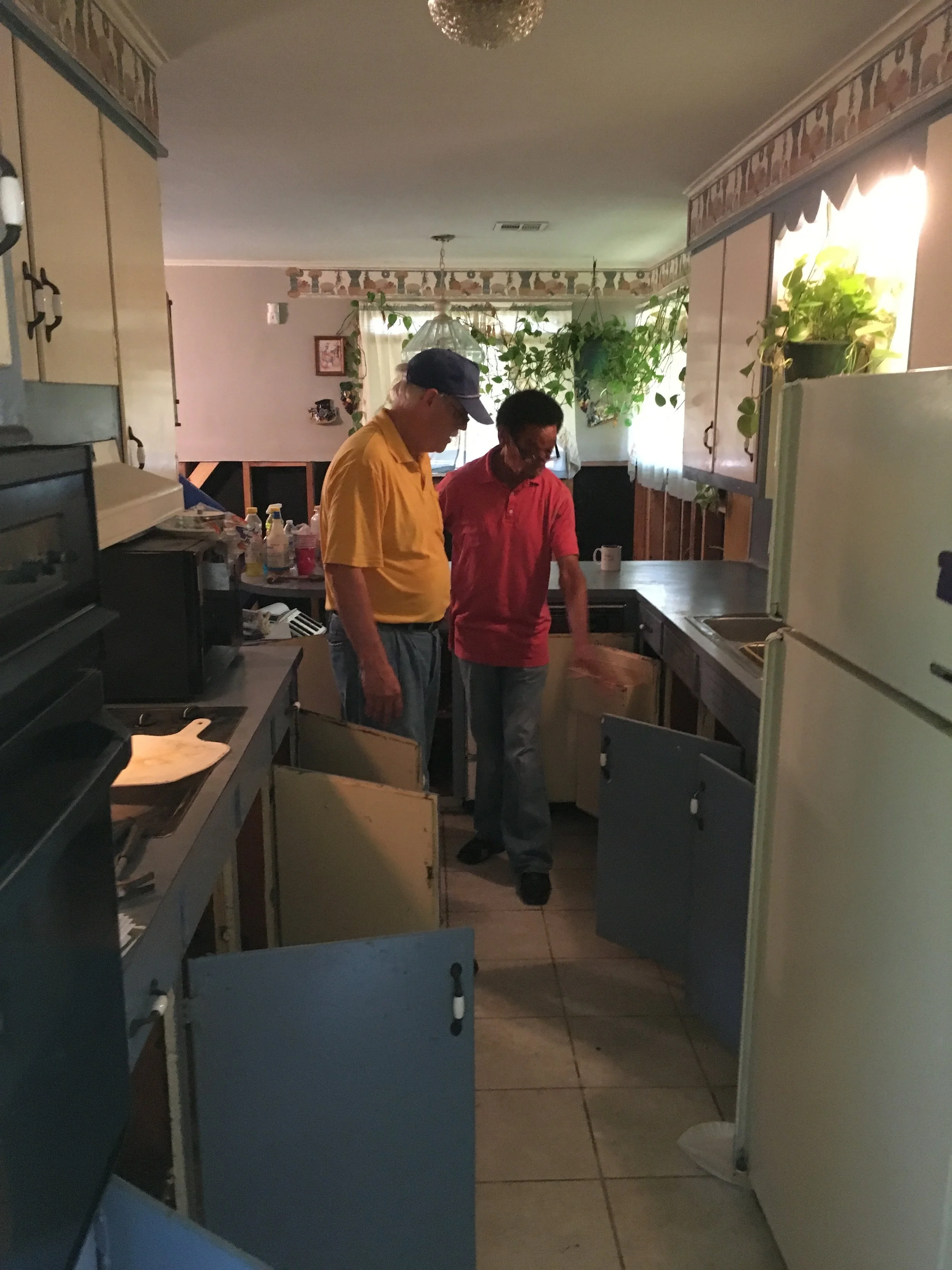 Two men standing in a kitchen inspecting open cabinet doors. One man is wearing a yellow shirt and a cap, and the other man is wearing a red shirt. The kitchen has plants on the window ledge and various items on the counter.