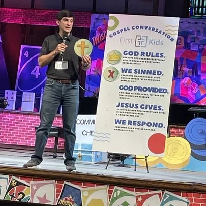 A young boy standing on a stage holding a microphone and a round sign with a cross. Behind him is a large whiteboard titled 'Gospel Conversation' with points about God, sin, Jesus, and response. The background features colorful decorations and banner