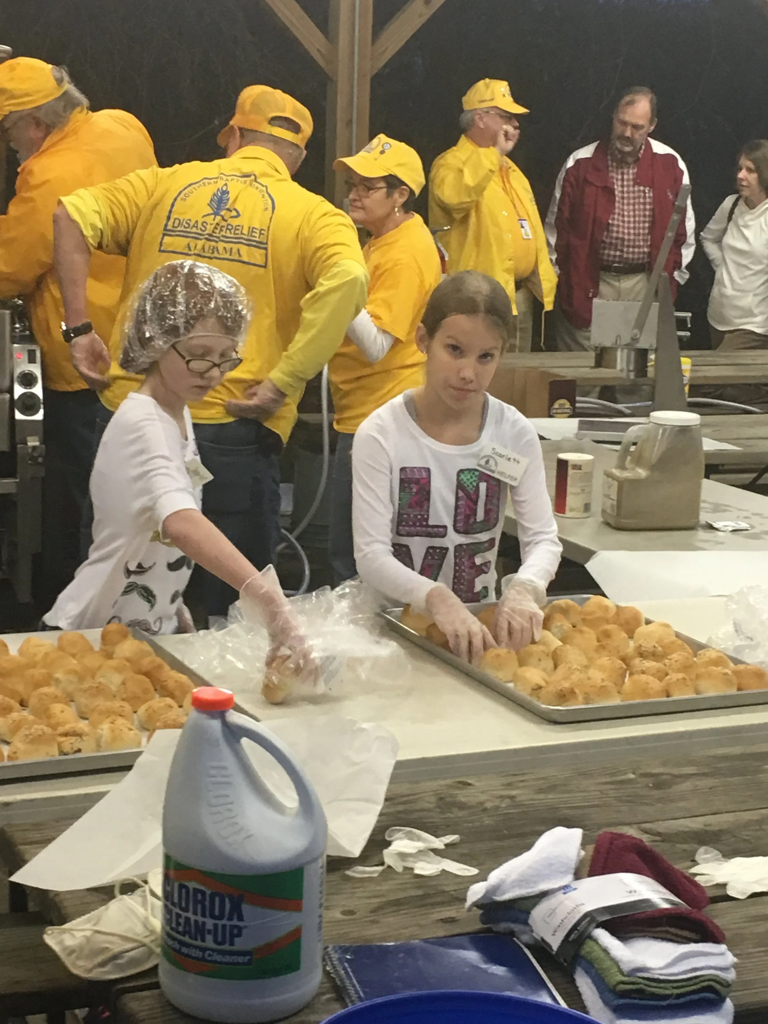 Two girls wearing gloves and working with baked goods at a baking event, with a group of adults wearing yellow shirts in the background.