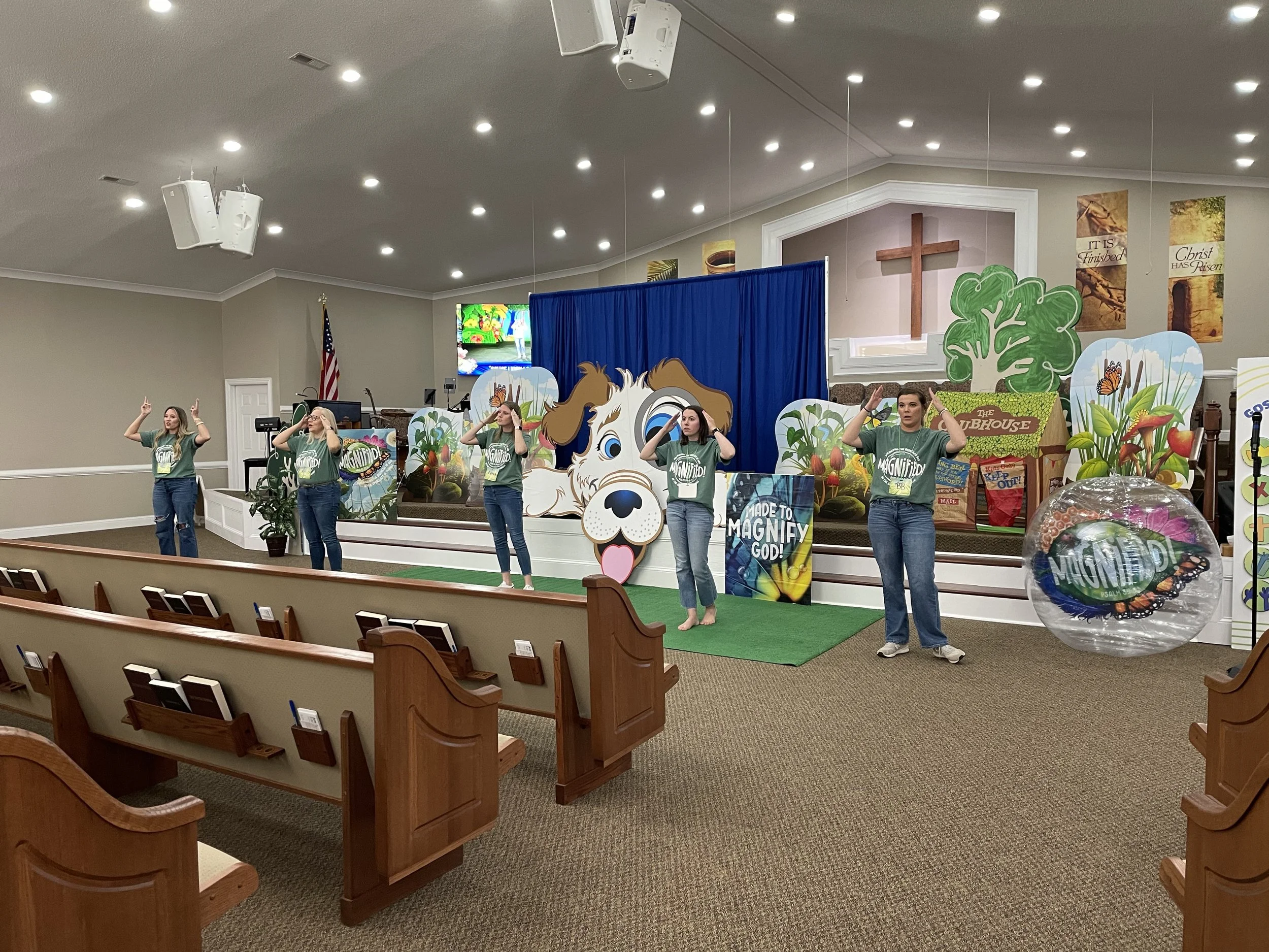 Women performing a song or dance on stage in a church setting with colorful decorations, banners, and a large dog cutout background.