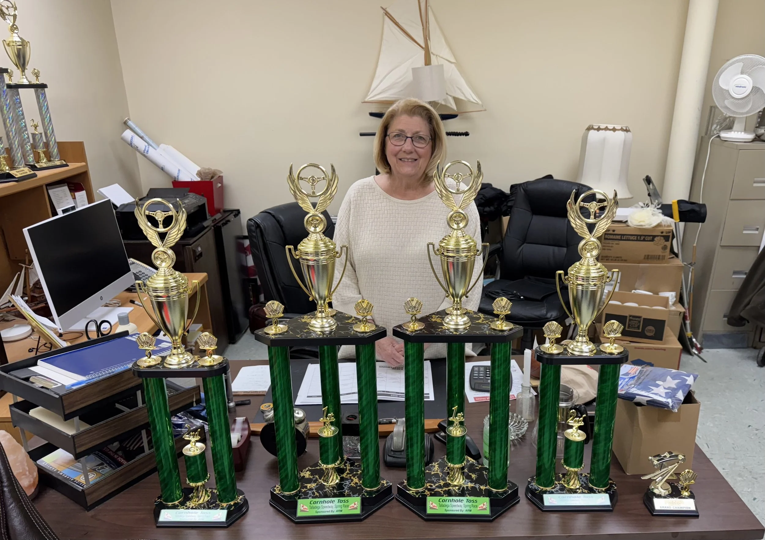 A woman standing behind a table with multiple large and small golden trophies, in an office room with desk items, a computer, and boxes in the background.