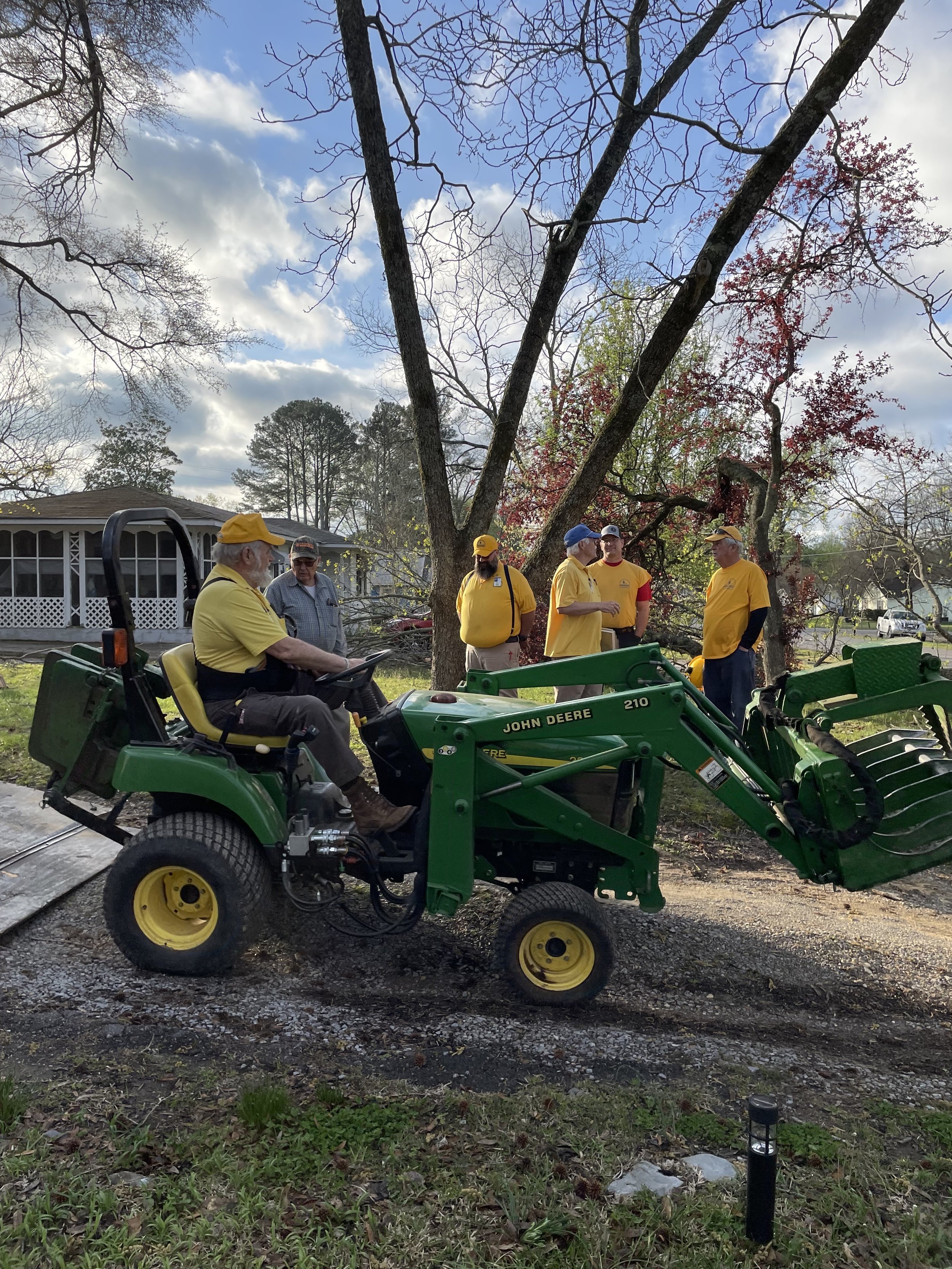 A group of five people wearing yellow shirts and hats are having a discussion outdoors near a large tree, while one person sits on a John Deere tractor.