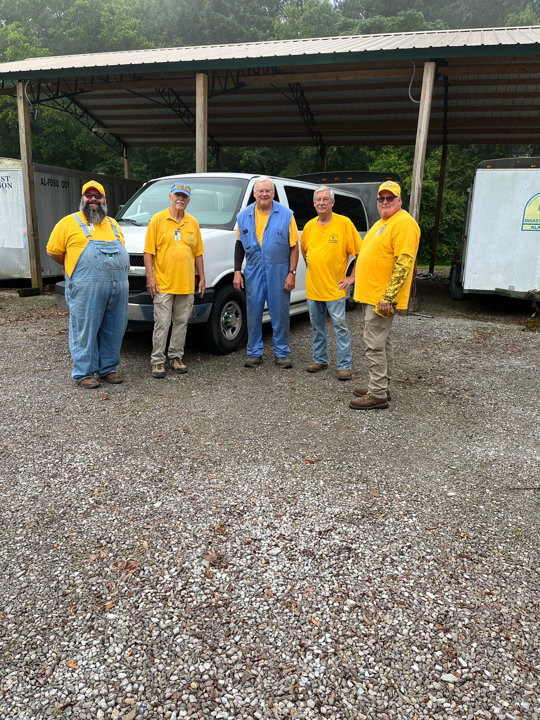 Group of five men standing in front of a white pickup truck under a metal shelter. Four men are wearing yellow shirts or jackets, and one is in blue. They are outdoors on a gravel surface, with trees visible in the background.