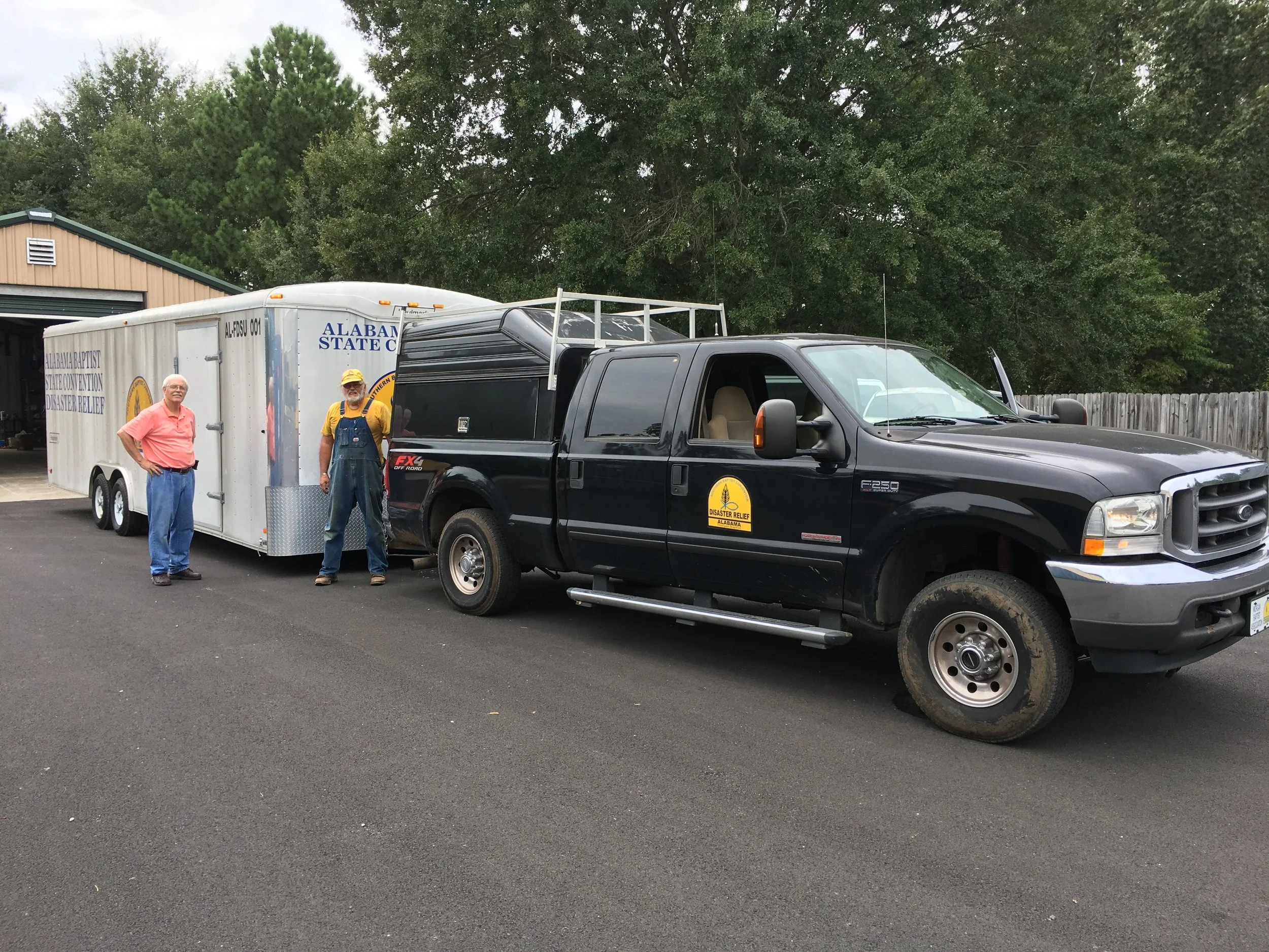 A black disaster relief truck parked on a paved surface next to a white trailer with a logo and text for Alabama State Fire and Rescue. Two men stand near the trailer, one wearing a pink shirt and the other in overalls and a yellow hat. A wooden fenc
