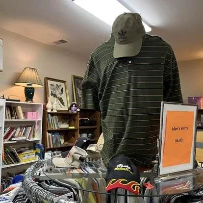 A clothing store display featuring a striped men's shirt on a mannequin with a beige cap, next to a sign that says 'Men's shirts $5.00',   surrounded by various hats, including a black cap with flames, and other apparel items.