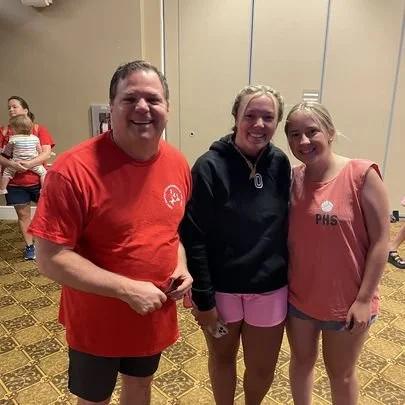 Three people standing together indoors, smiling at the camera. The man on the left is wearing a red shirt and black shorts. The woman in the middle is wearing a dark hoodie and pink shorts. The young girl on the right is wearing a pink shirt and pink