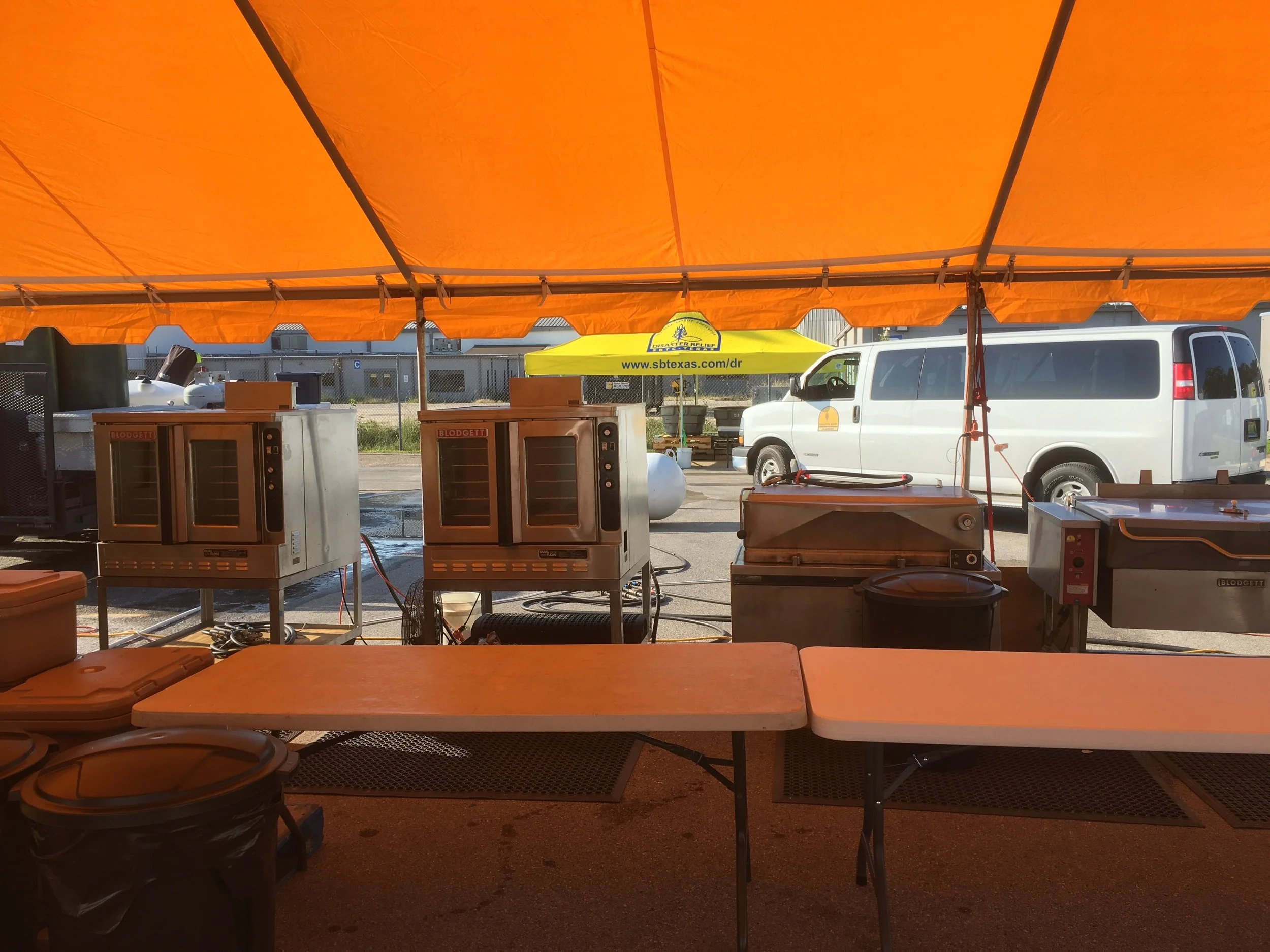 A view of a food stall setup under an orange canopy, with two large food warmers, a cooking griddle, and tables in the foreground. A white van and a yellow tent can be seen in the background.