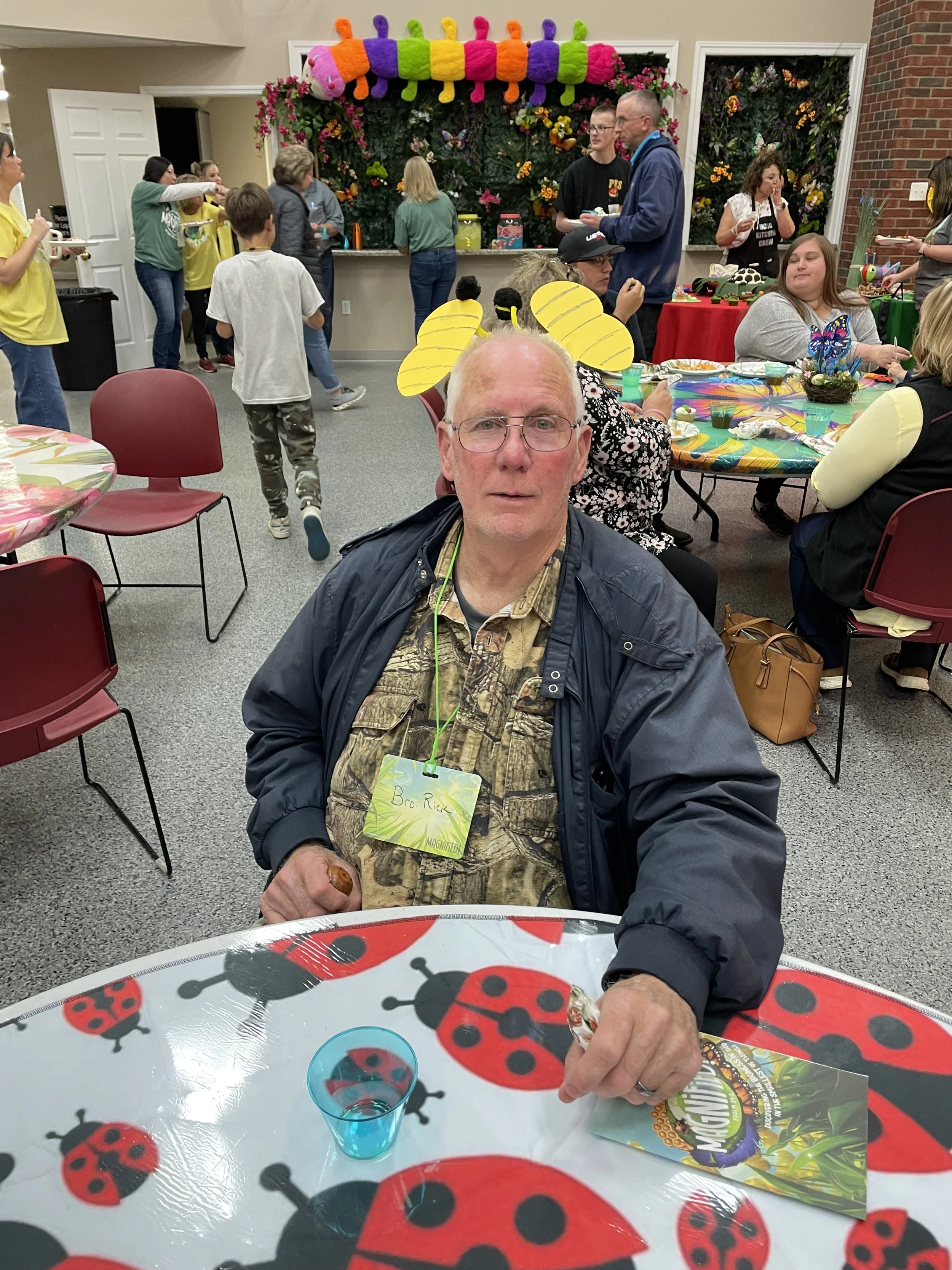 A man with glasses and a yellow headband with bee antennae sitting at a table decorated for a party, with a ladybug-themed tablecloth, a blue drink, and a menu, in a room filled with people, some dressed in yellow, engaging at a celebration.