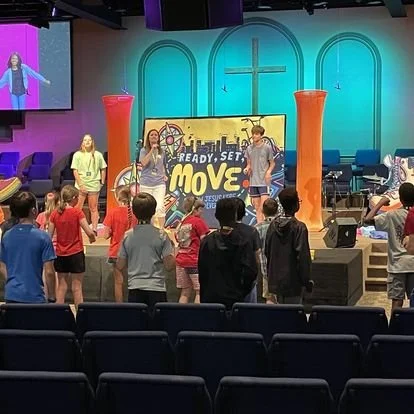 Children participating in a Move! fitness class led by instructors in a church or community center with a colorful stage, large orange pillars, and a backdrop featuring a cityscape and the word 'Move'.