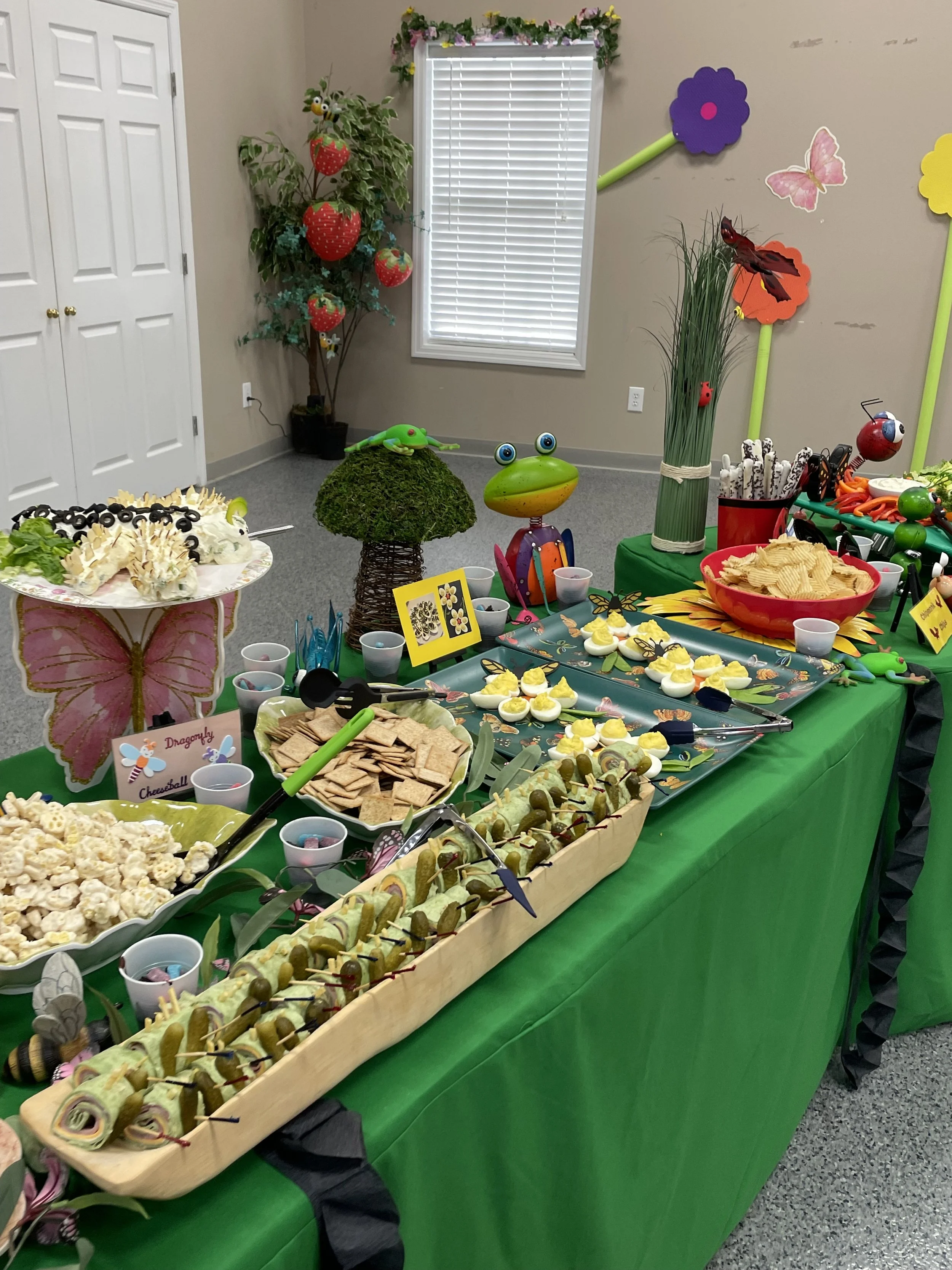 Colorful birthday party table with snacks and decorations, featuring frog and butterfly themed decor, in a room with plants and butterfly wall art.