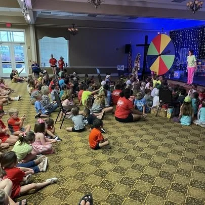 Children gathered in a room watching a live performance on stage with a multicolored spinning wheel and a blue backdrop.