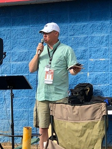 A man wearing a white cap and sunglasses stands outdoors, speaking into a microphone, with a blue wall behind him and a beige and green stroller nearby.