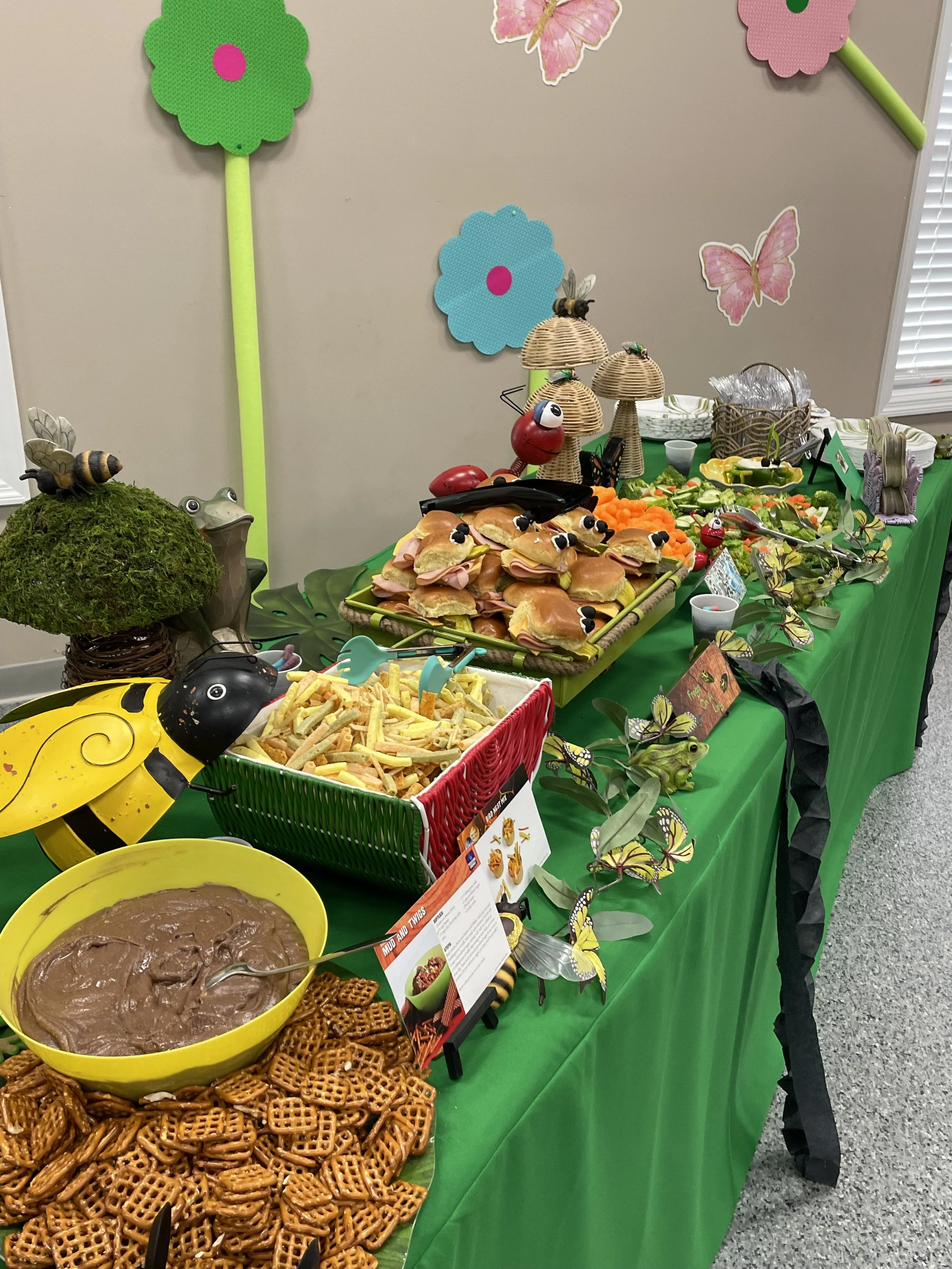 A table decorated with a bug and butterfly theme, displaying various snacks such as sandwiches, chips, and dip, with paper flowers and butterfly decorations on the wall.