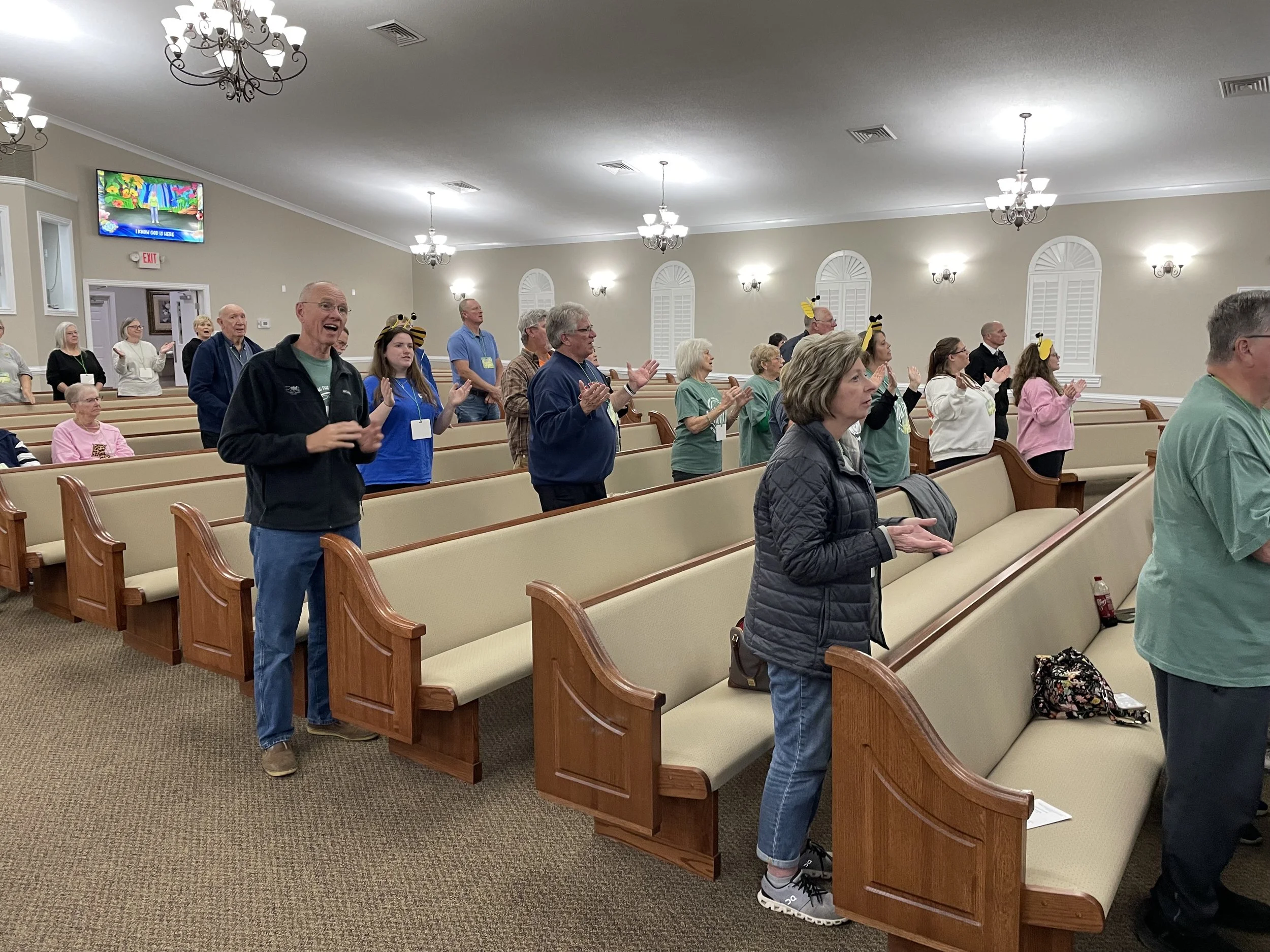 Group of people standing and clapping in a church or similar setting, some wearing bee headbands, with empty pews, chandeliers, and a television screen in the background.