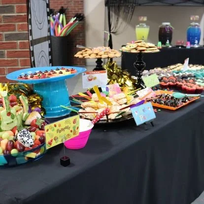 A table decorated for a celebration with various colorful treats, including cupcakes, cookies, and candy in bowls and plates, with beverage bottles and a backdrop of colorful jars in the background.
