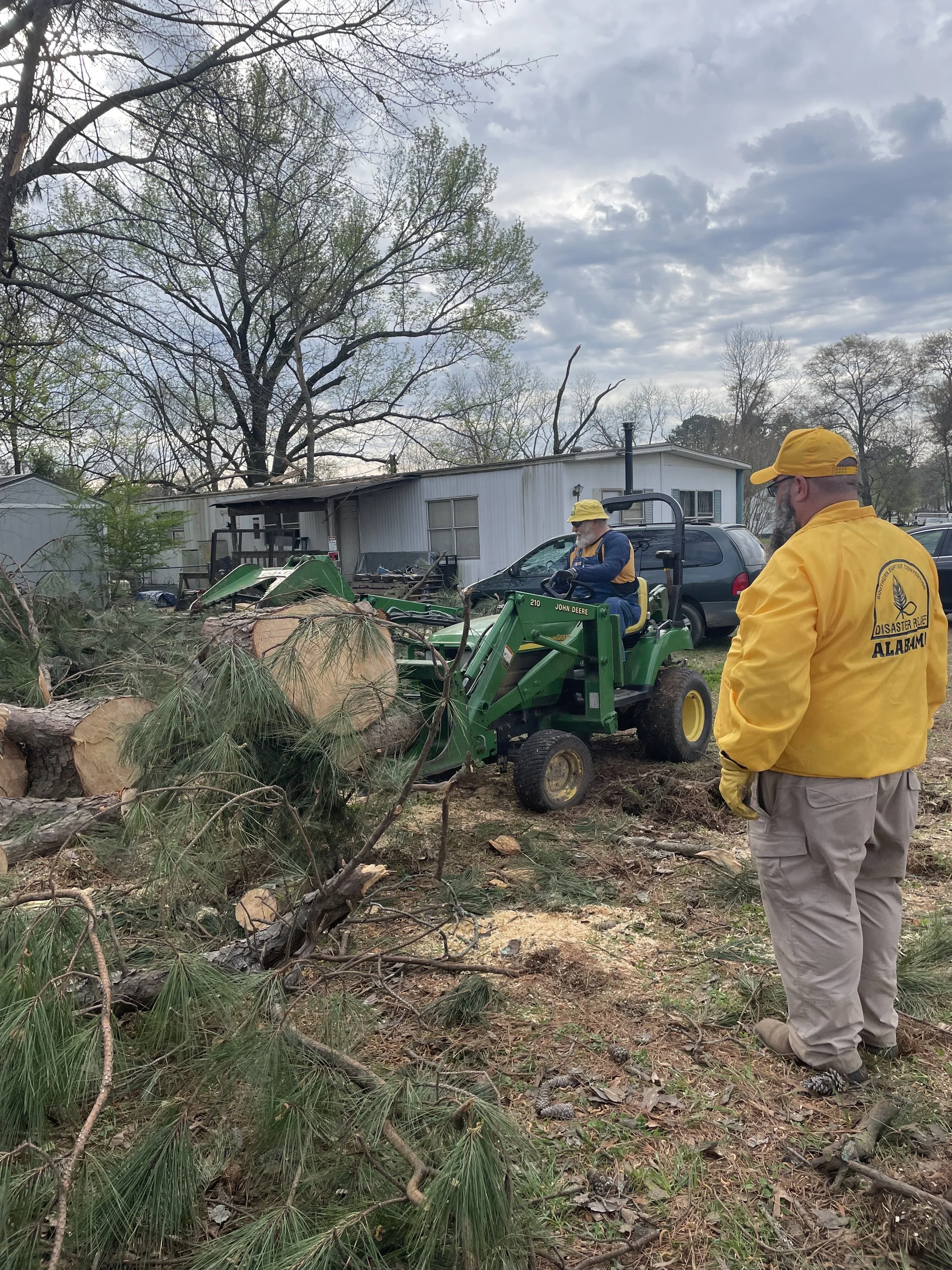 Workers clearing fallen trees with a small John Deere tractor and wood chips on the ground in a residential area.