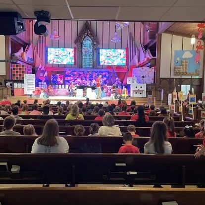 Audience members seated in a church auditorium watching a colorful stage performance with digital screens and a stained glass window behind.