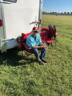 Man sitting on red folding chair outside near a white trailer with a grassy field in the background.