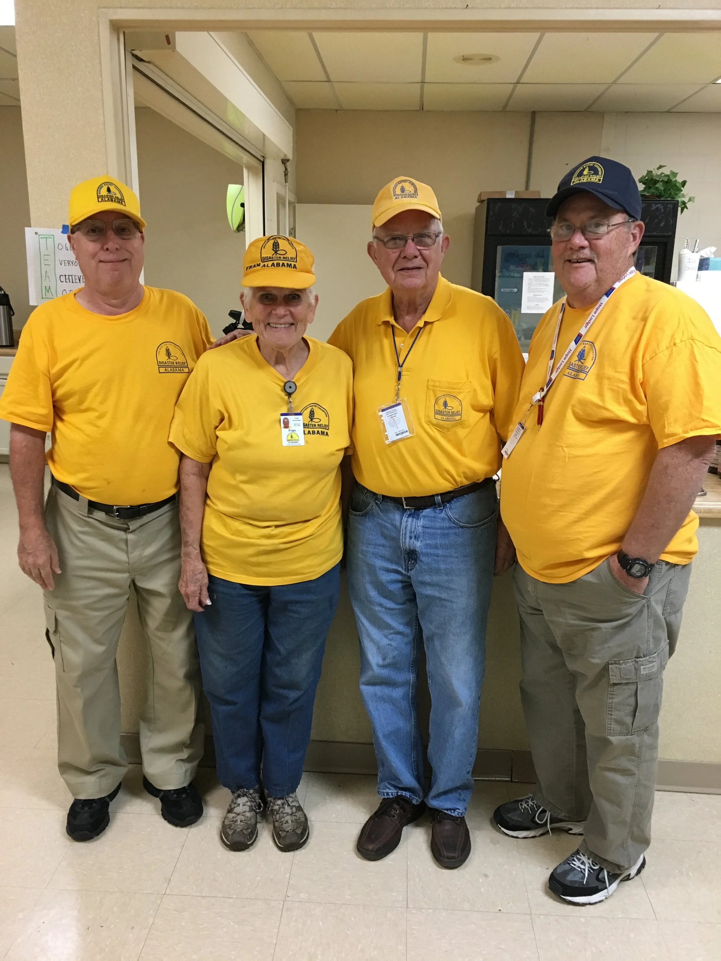Group of four elderly volunteers wearing yellow 'Disaster Relief Alabama' shirts and caps, standing together in an indoor setting.