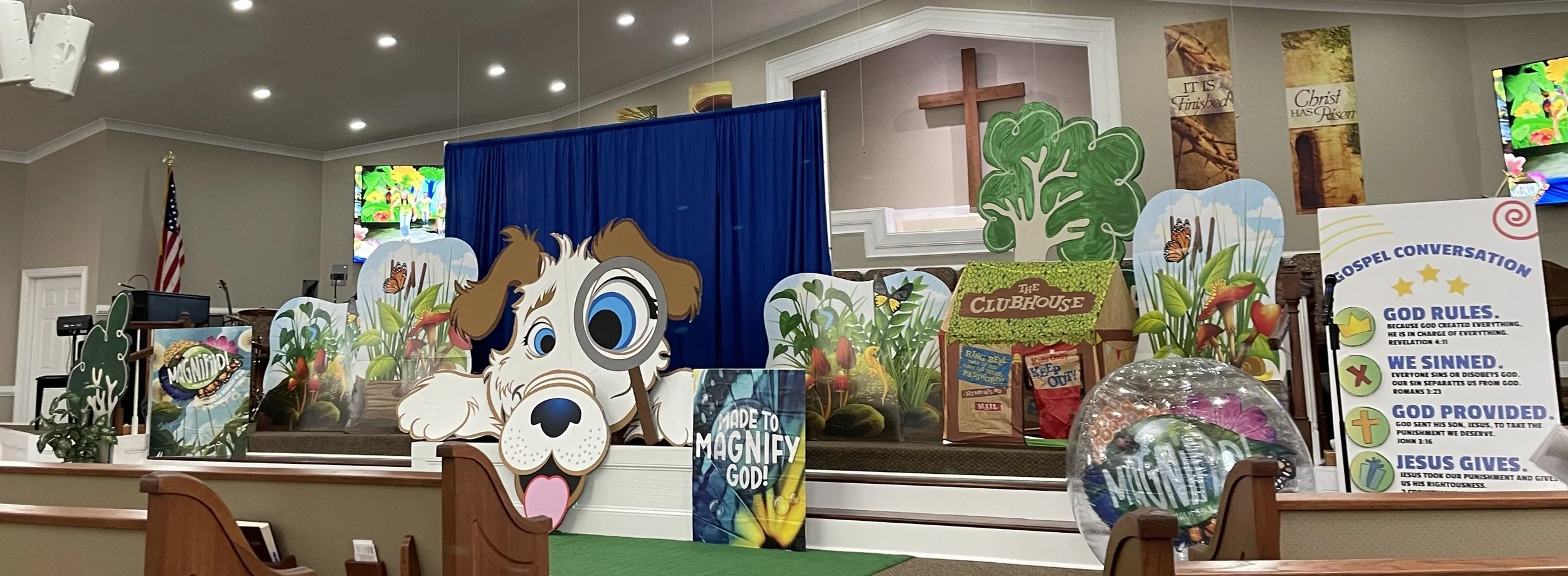 Stage decorated with Christian and nature-themed cutouts, including a cartoon dog with a monocle, a cross, a large green tree, and signs with religious messages in a church setting.