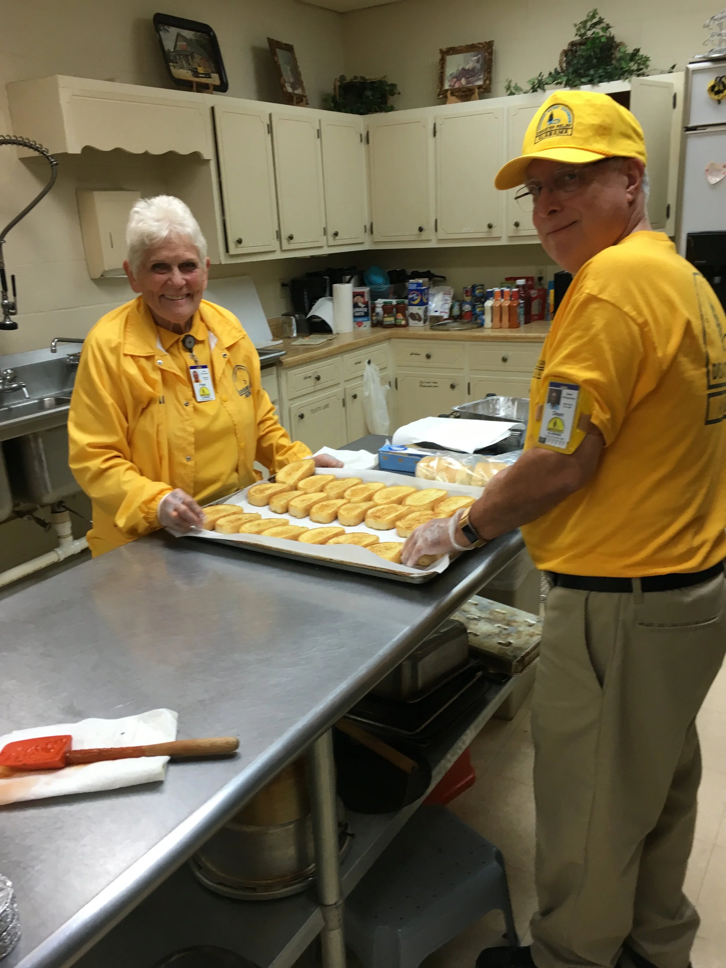 Two older adults dressed in yellow uniforms preparing sandwiches on a tray in a kitchen.