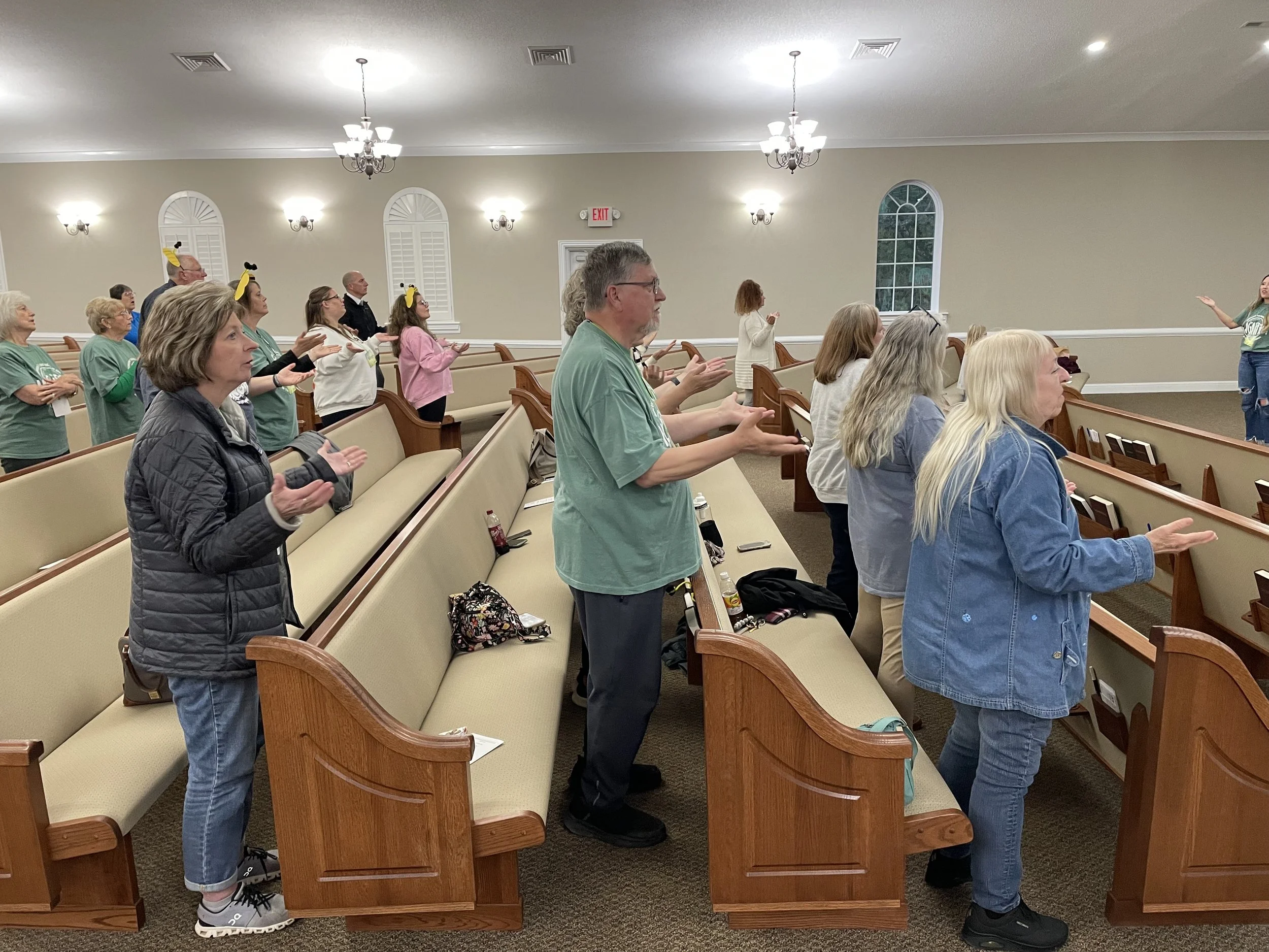 People standing in pews inside a church, engaged in prayer or worship, with some wearing yellow headbands, and a woman addressing the congregation from the front.