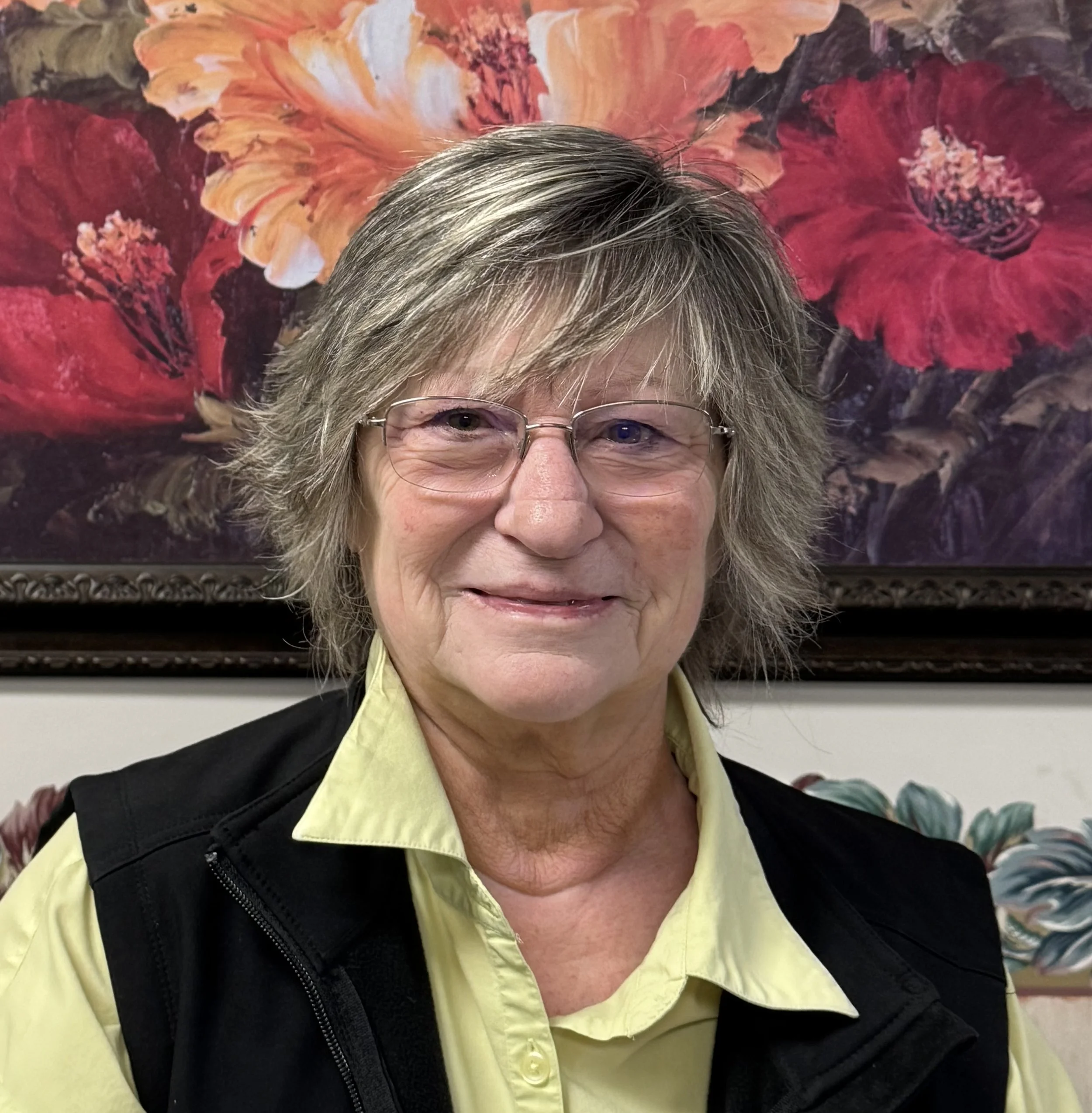 A woman with gray, wavy hair and glasses, smiling, wearing a yellow shirt and a black vest, in front of a colorful floral painting featuring large red and orange flowers.