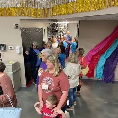 Group of people, including children and adults, gathered in a decorated indoor space with colorful streamers and gold fringe decorations, likely for a celebration or party.
